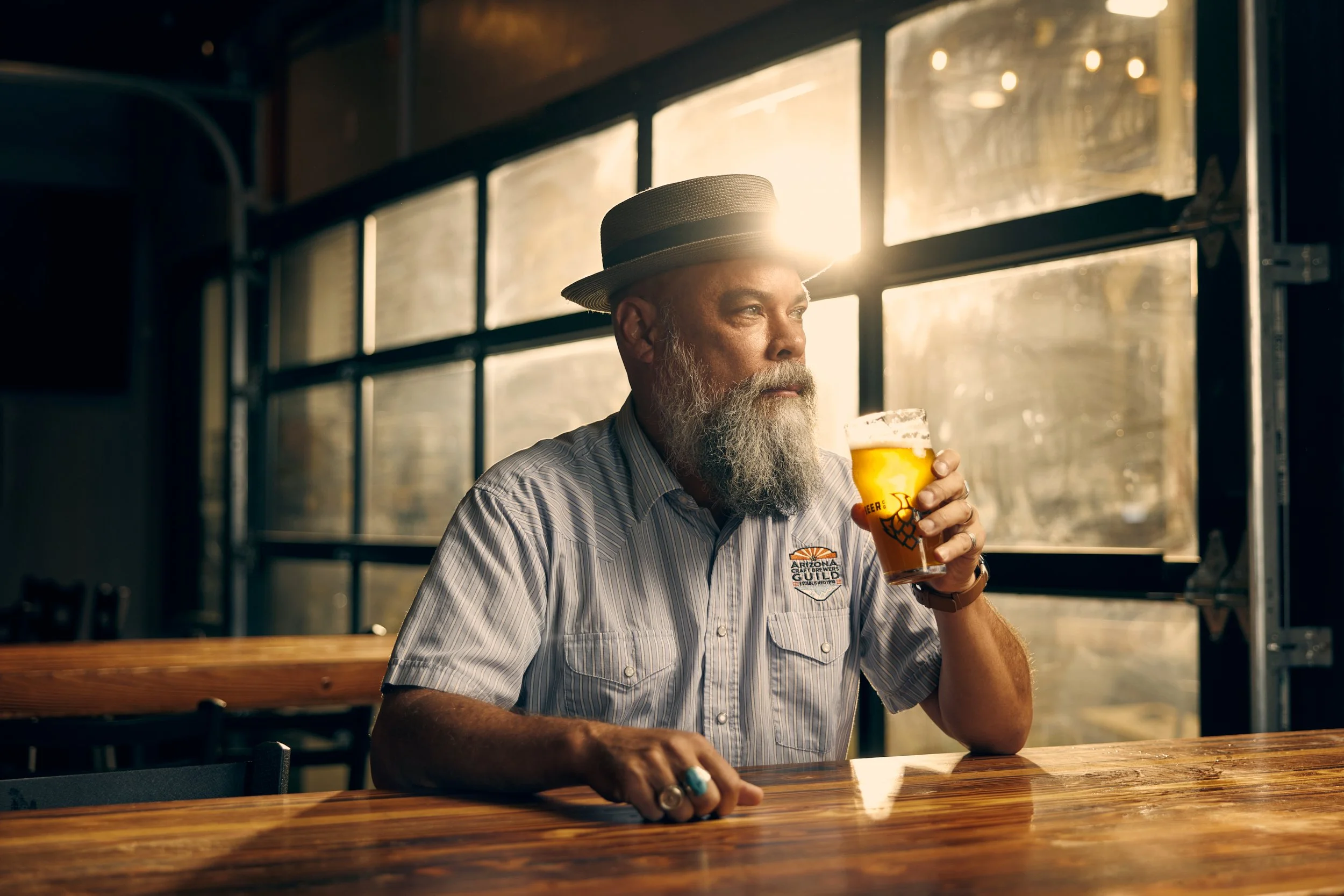 Older man with a beard wearing a straw hat and striped shirt sitting at a wooden bar, holding a glass of beer, with sunlight streaming through large windows behind him.