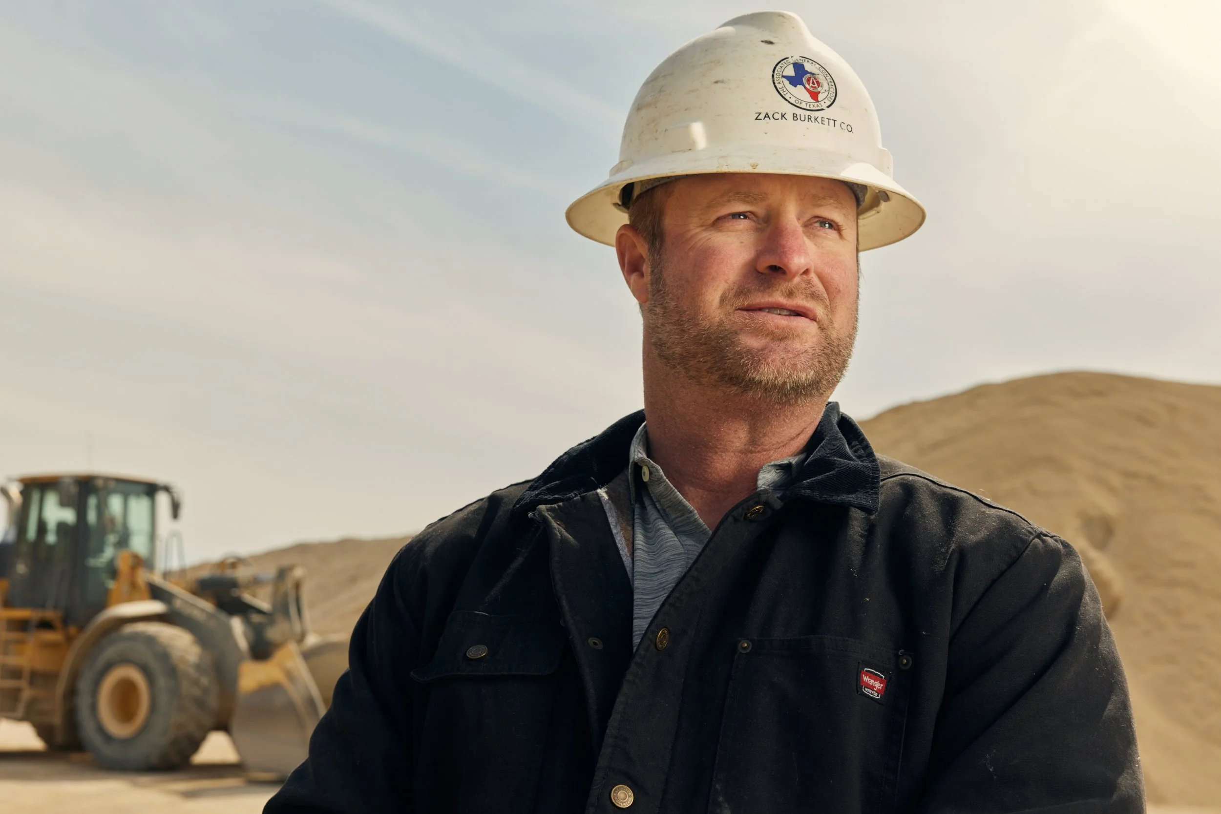 A man wearing a white construction helmet and a black jacket standing outdoors with construction equipment and a hilly landscape in the background.