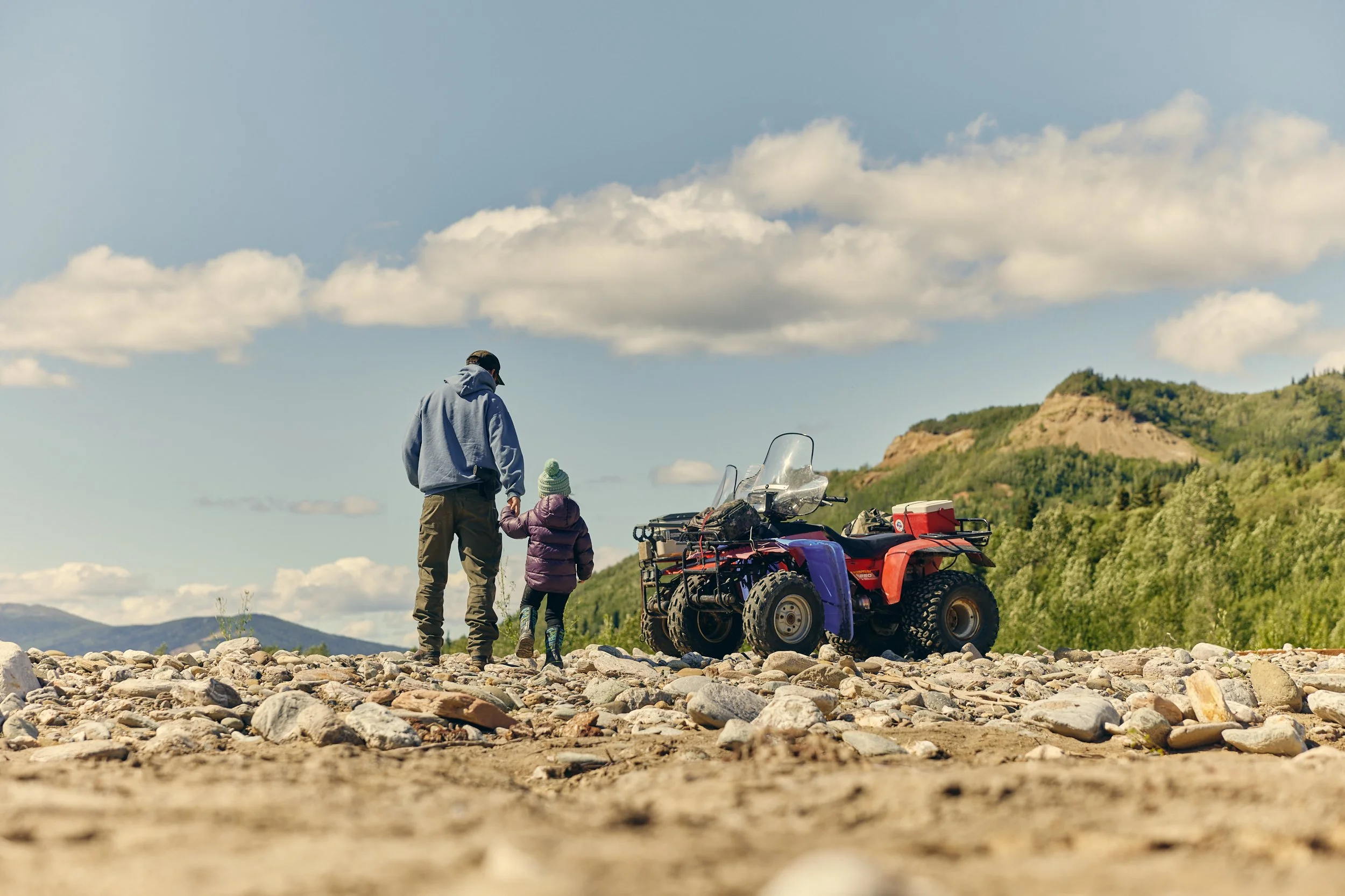 A man and a young girl walking on rocky ground near a red ATV with gear, set against green hills and a blue sky with clouds.