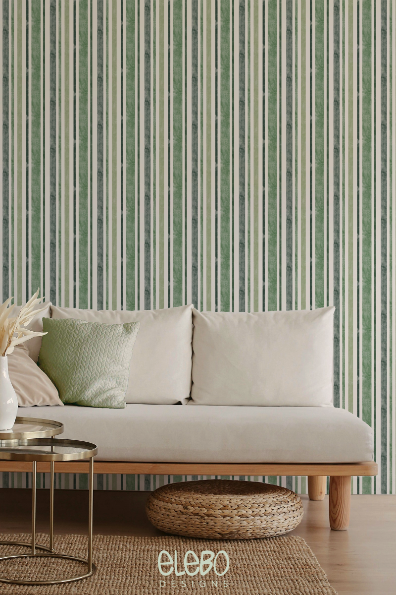 Vertical striped traditional non-pasted wallpaper in forest and sage green tones with a subtle watercolour texture, styled in a cozy living room setting featuring a low-boho sofa, light wood furniture, and natural woven accents.