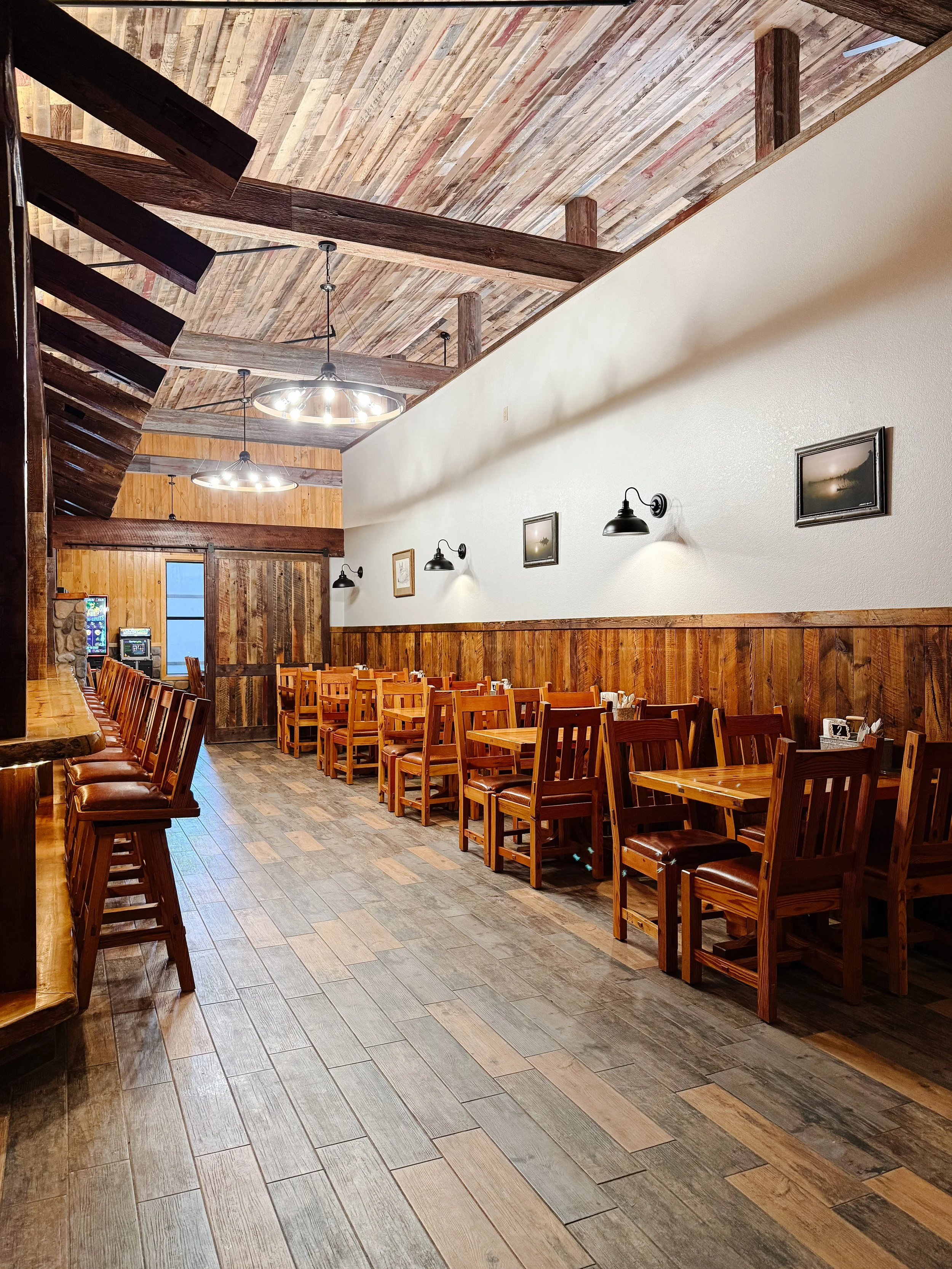 Interior of a rustic bar and restaurant with wooden tables and chairs, a long stone bar counter, and festive holiday decorations including lights and garlands.