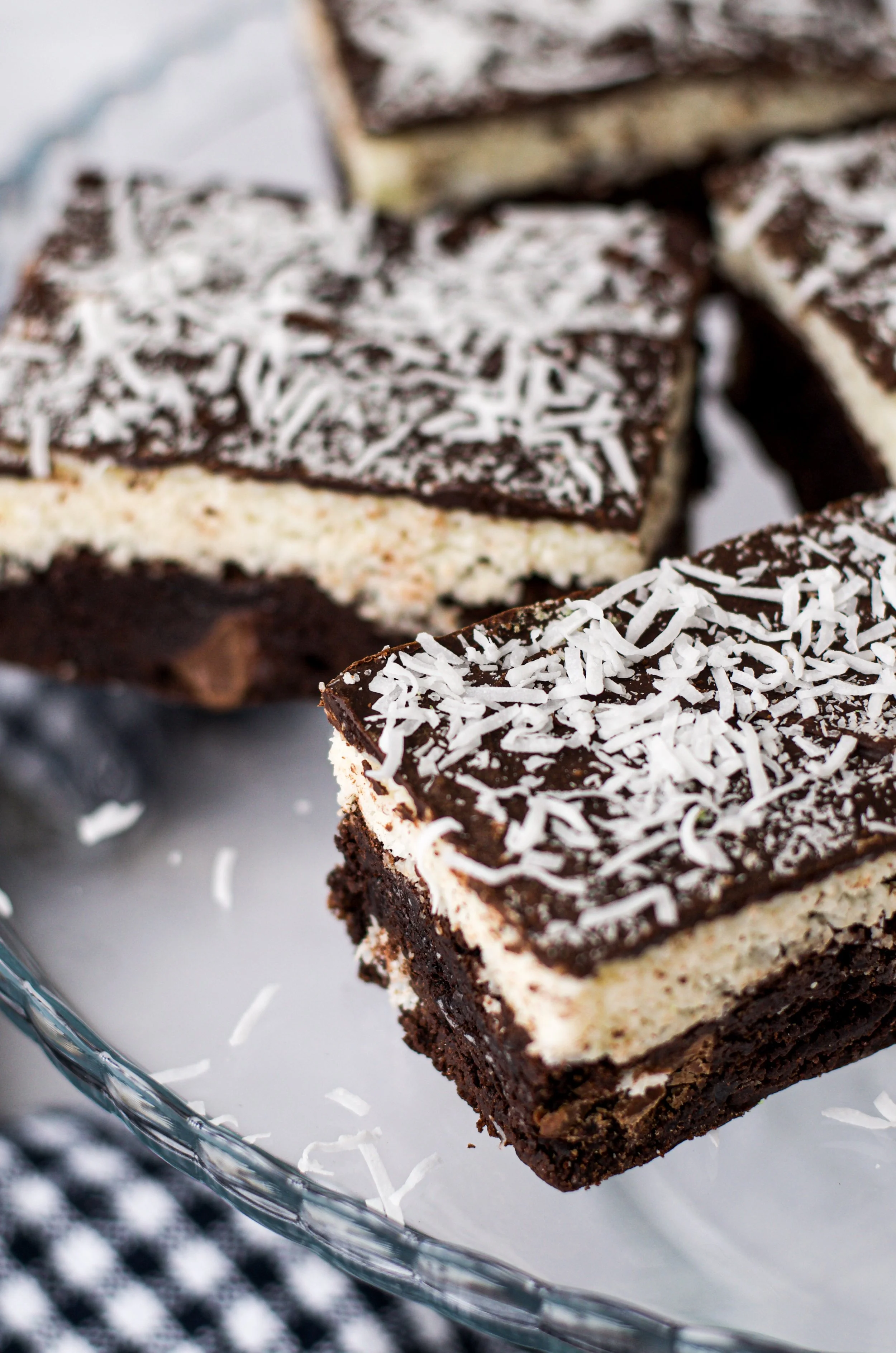 Close-up of chocolate and vanilla layered dessert squares topped with shredded coconut on a glass serving tray with a blurred checkered cloth underneath.