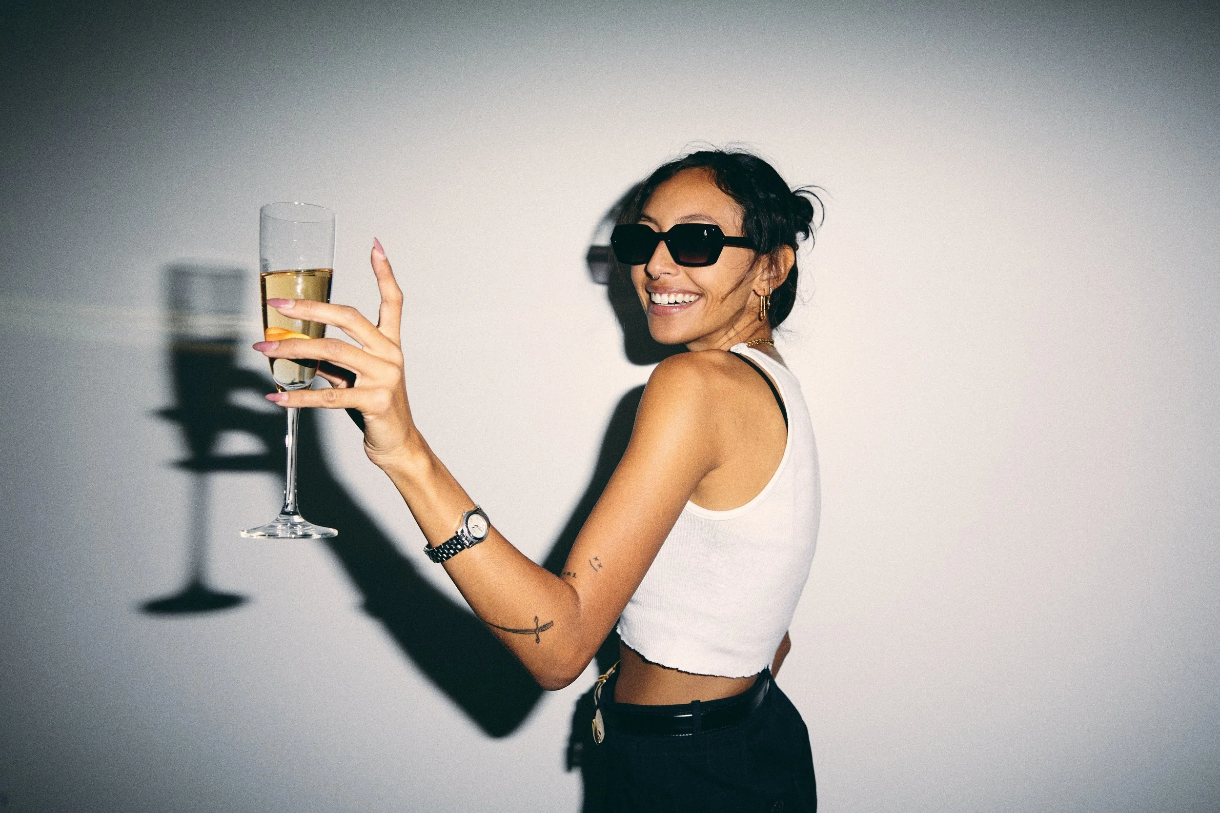 Woman in sunglasses holding a champagne flute, wearing a white tank top, smiling at camera.