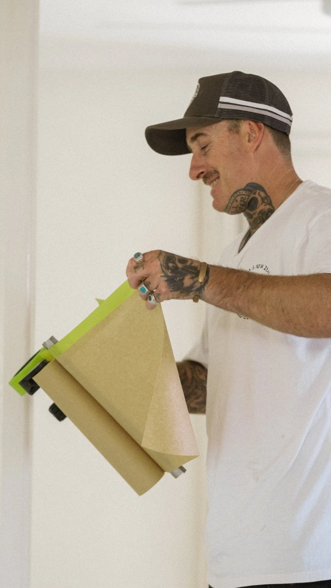 Person using a masking tool with paper, wearing a cap and white shirt, showing tattoos on arm and neck.