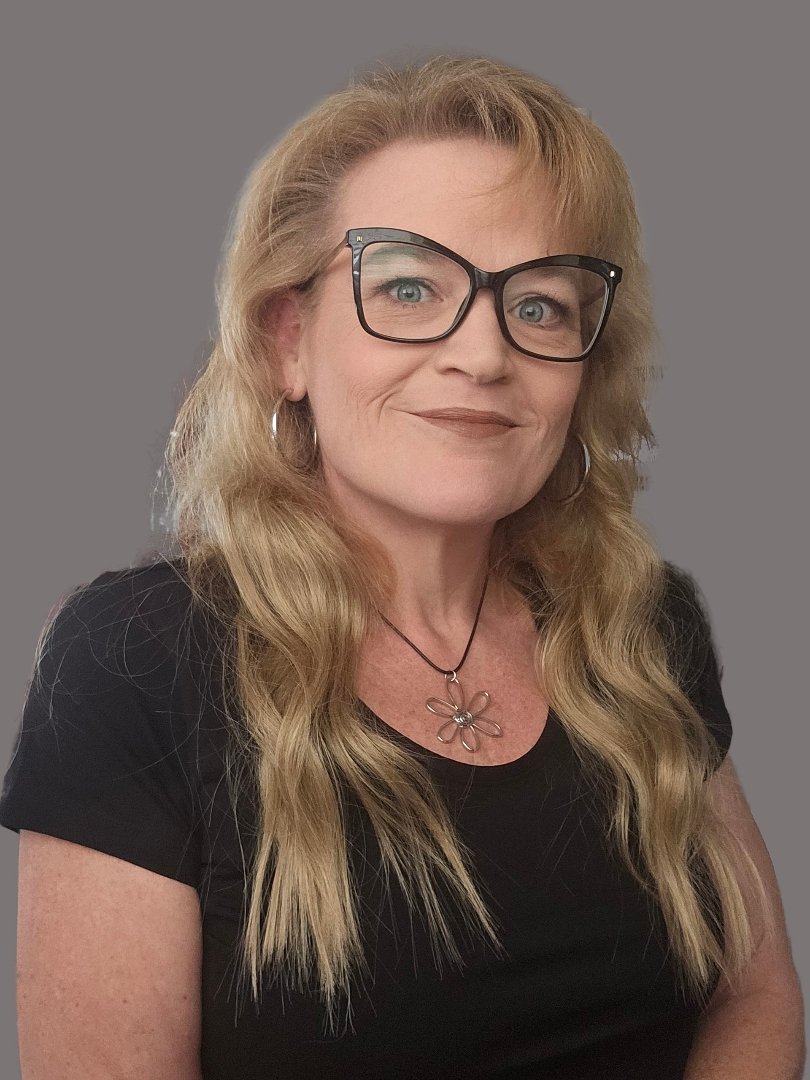 Woman with long wavy blonde hair, wearing black glasses, black top, and a flower pendant necklace, against a gray background.