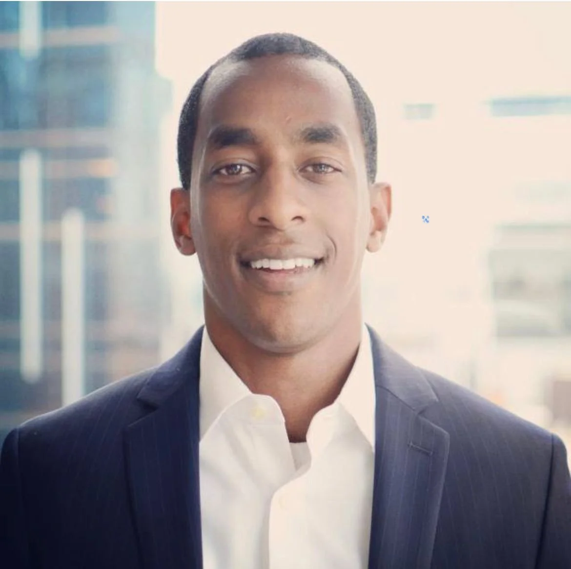 Smiling person in a business suit with a blurred cityscape background.