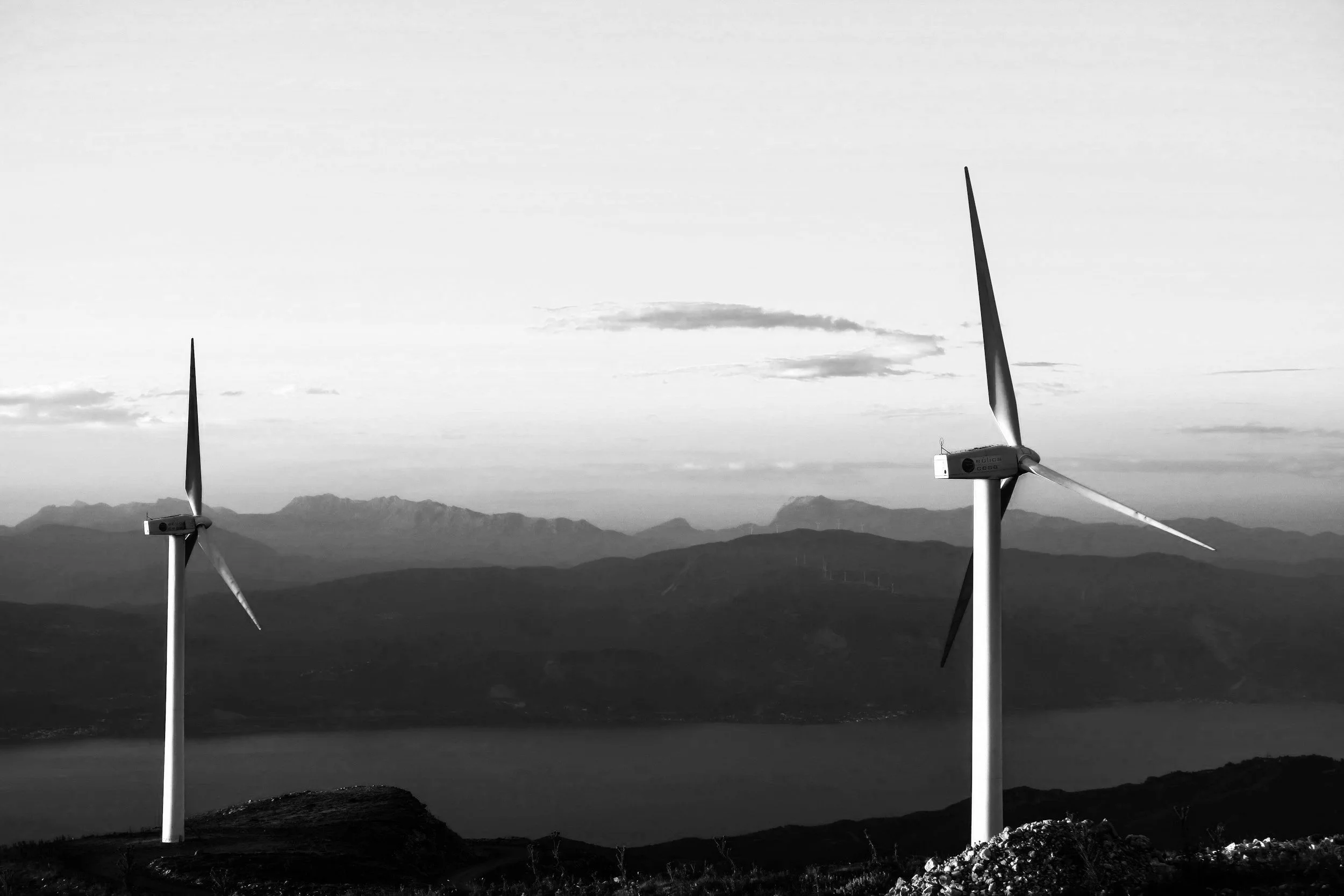 Black and white photo of three wind turbines on a hilltop with mountains and a cloudy sky in the background.
