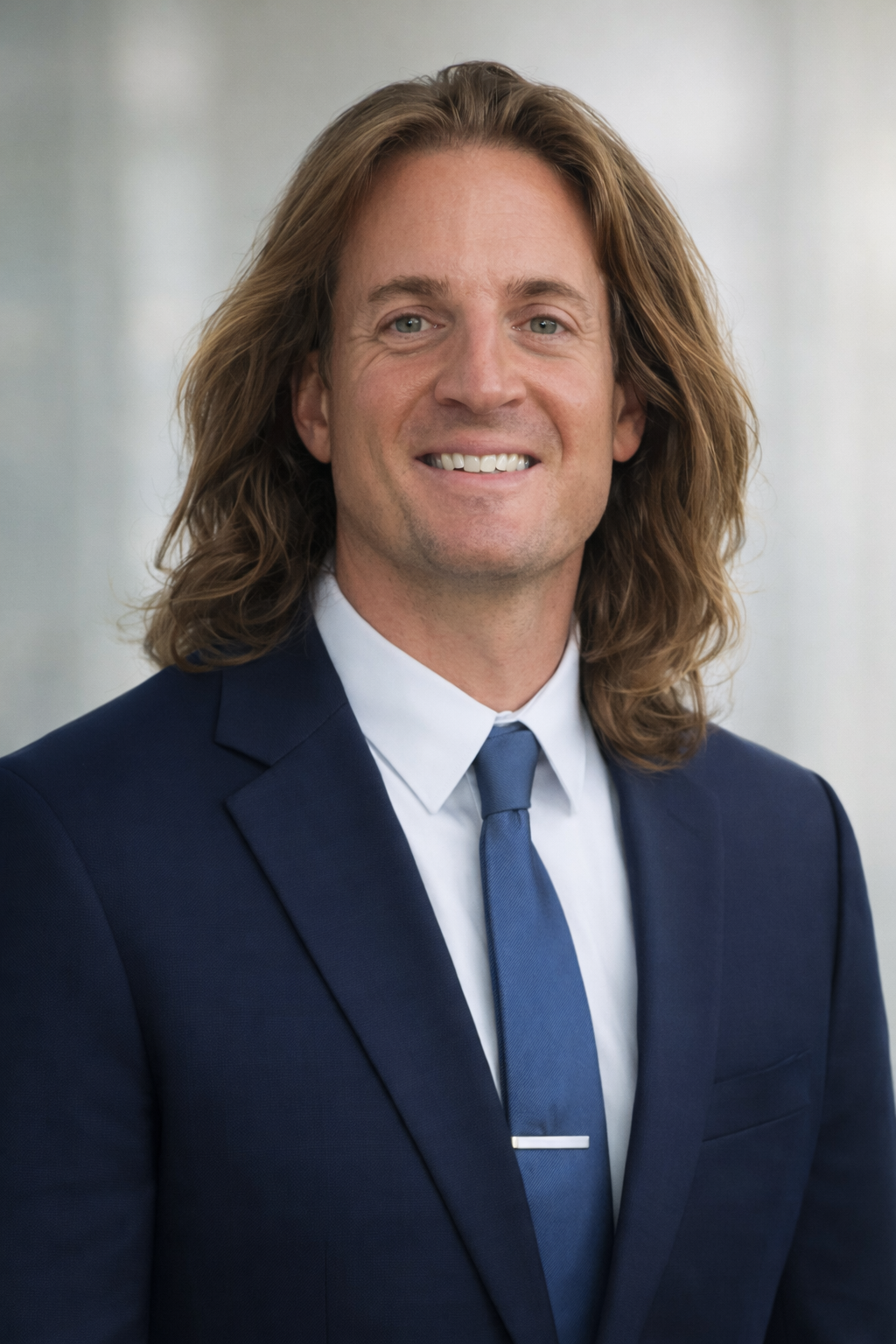 A man with long, wavy brown hair, wearing a navy blue suit, white shirt, and a blue tie, smiling against a blurred indoor background.