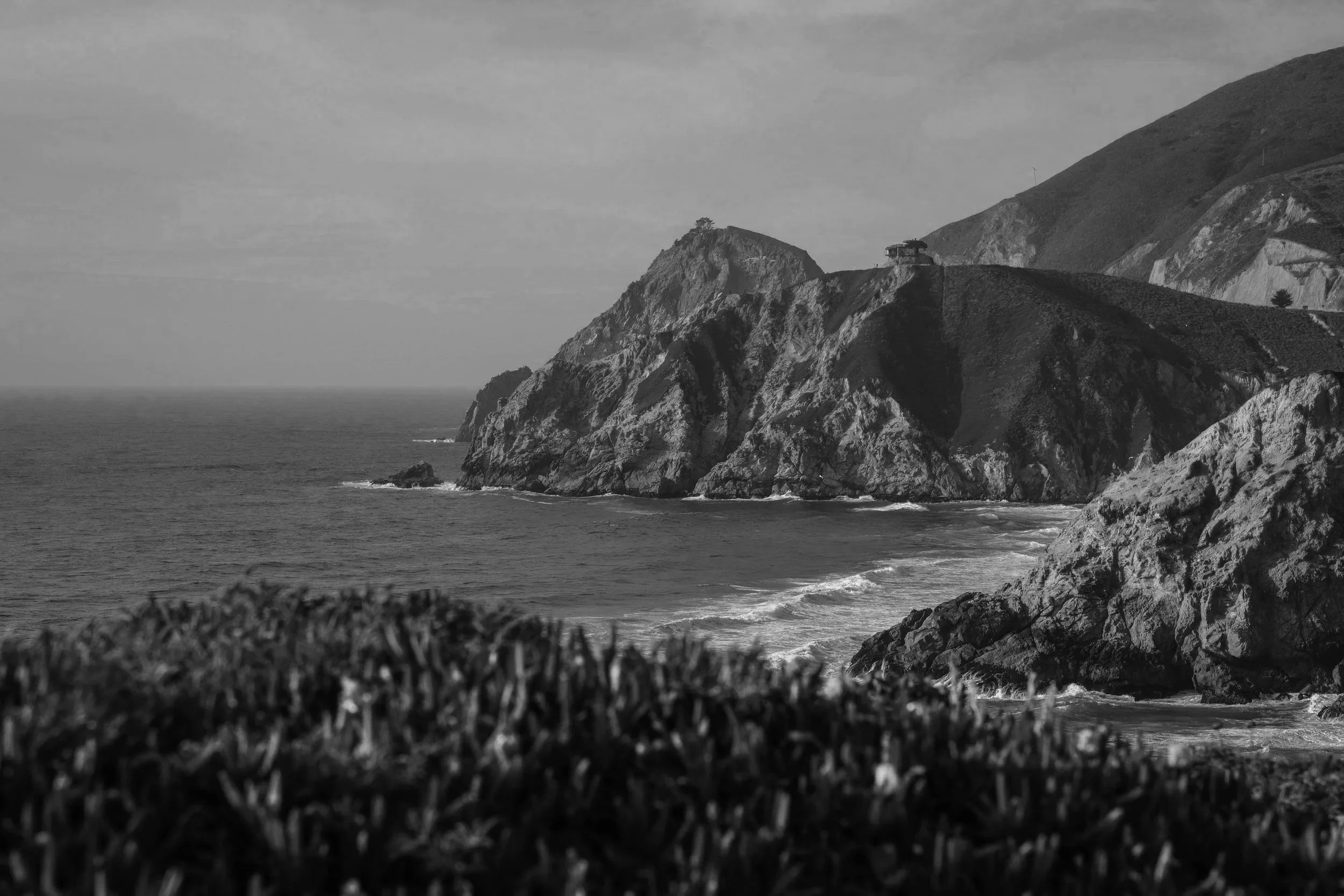 Black and white photo of a coastal cliff with buildings on top, overlooking the ocean with waves, grassy foreground, cloudy sky, and rugged terrain.
