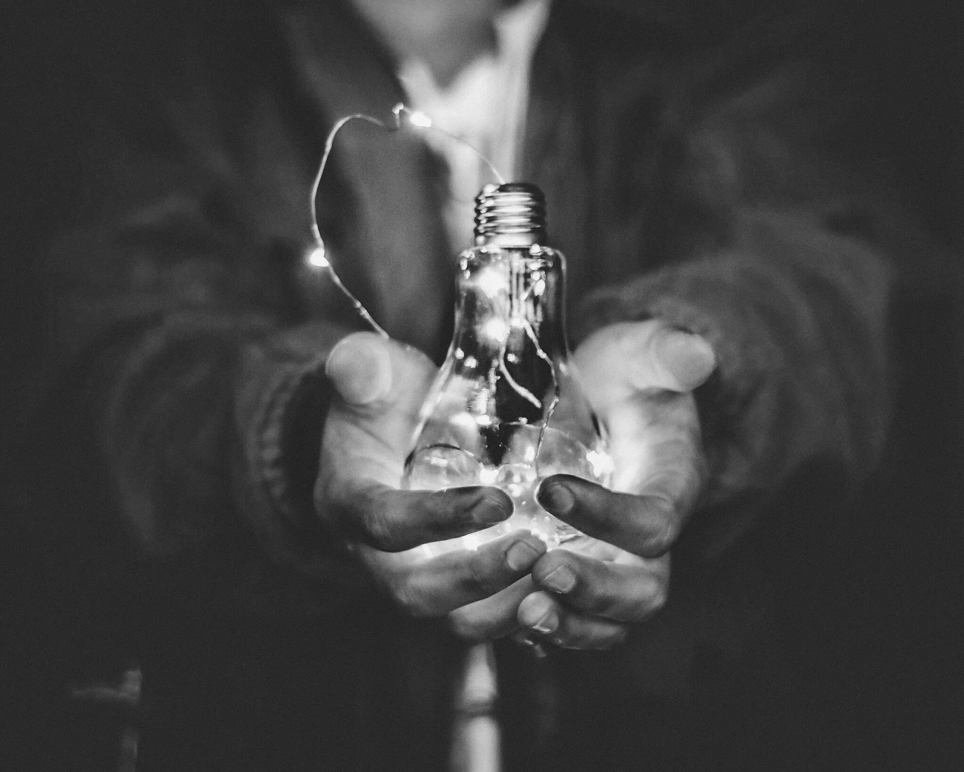 Black and white photo of hands presenting an illuminated lightbulb of ideas.