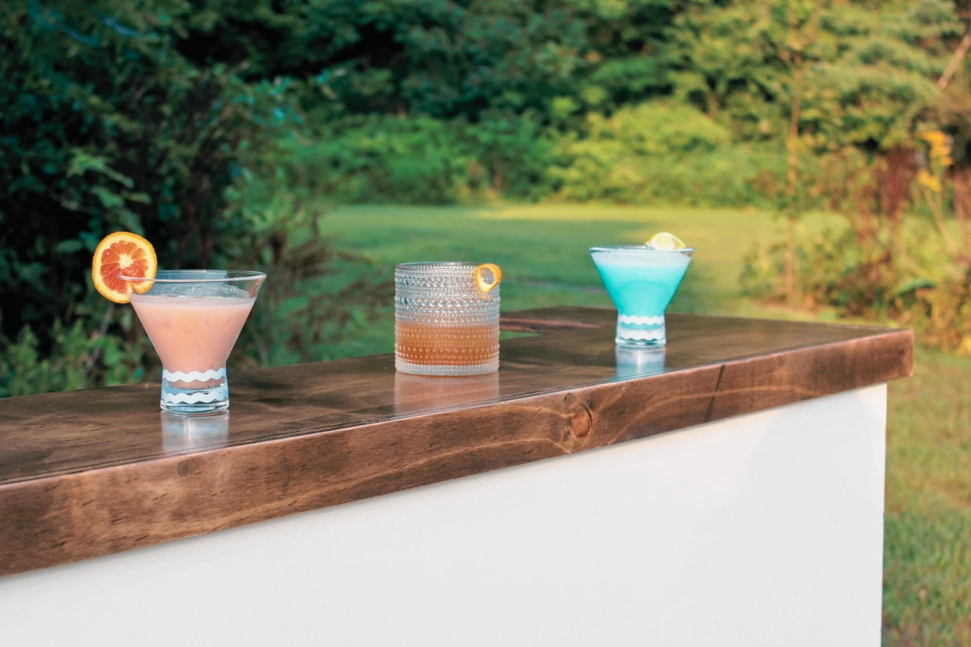 Three colorful cocktails on a wooden bar counter outdoors with green trees and grass in the background.