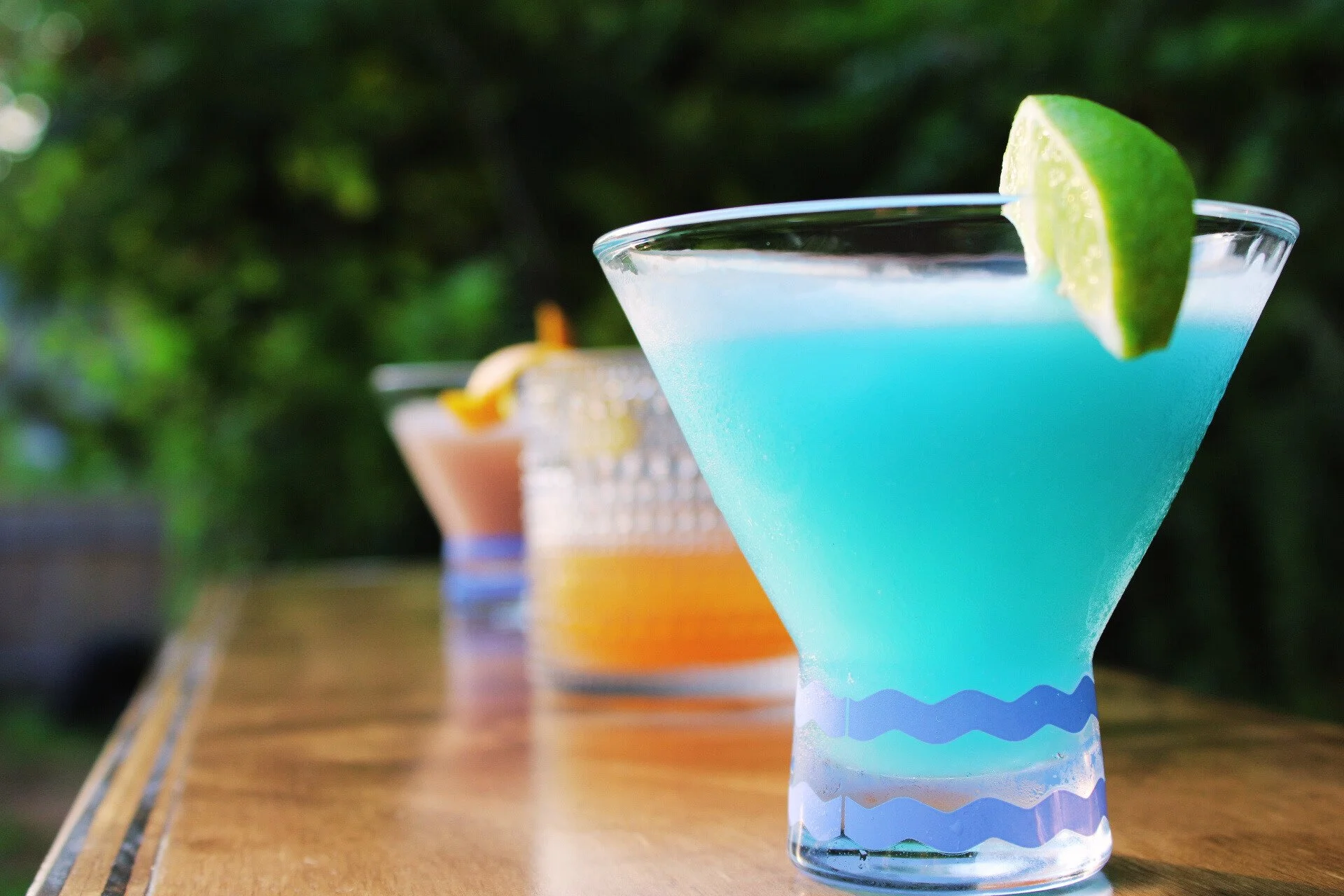 A close-up of a blue cocktail with a lime wedge garnish on a wooden bar or table, with two other colorful drinks blurred in the background against a green outdoor setting.