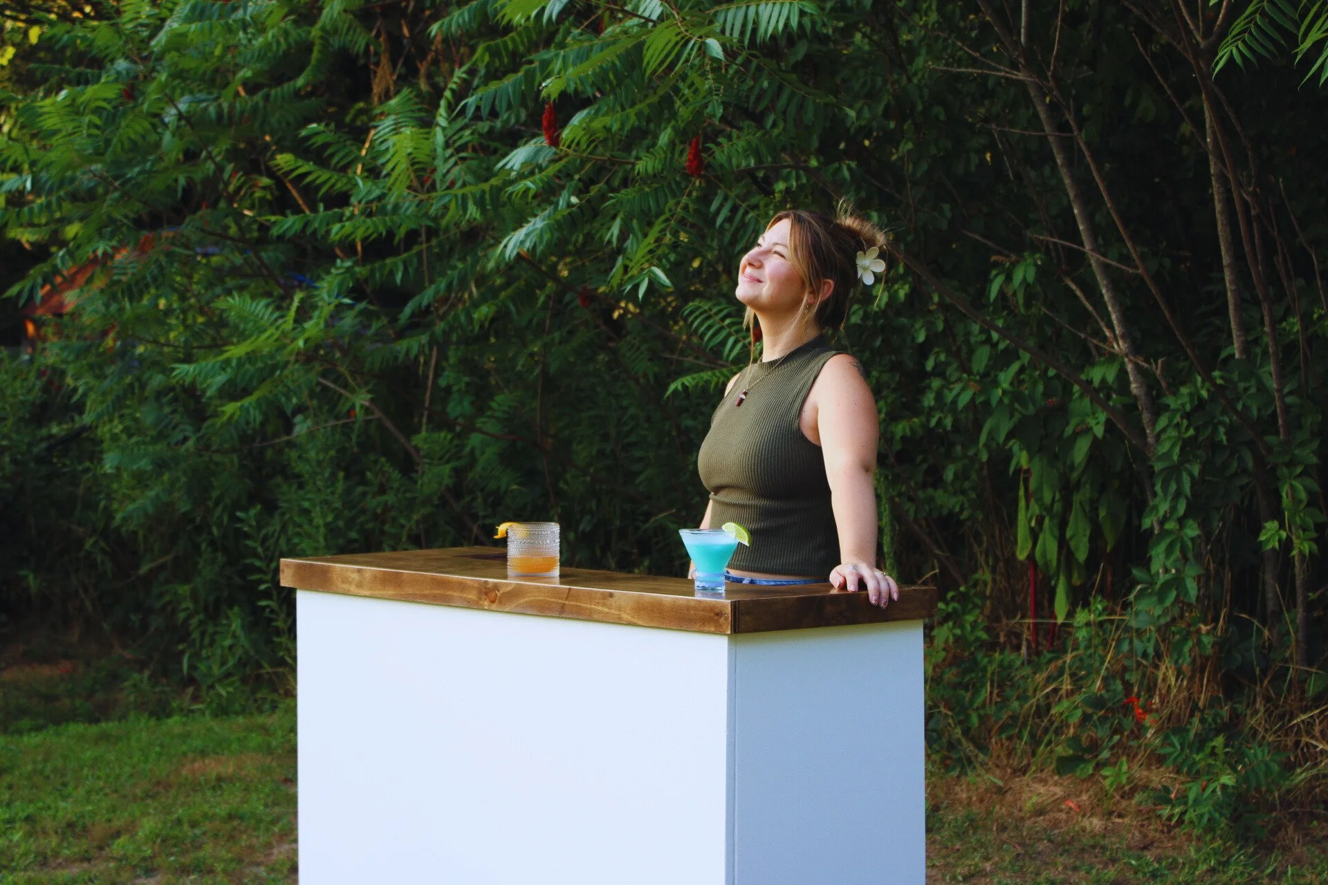 A woman with a white flower in her hair, wearing a sleeveless green top, stands behind a white bar with a wooden top, smiling with her eyes closed in a lush green outdoor setting with trees and foliage.