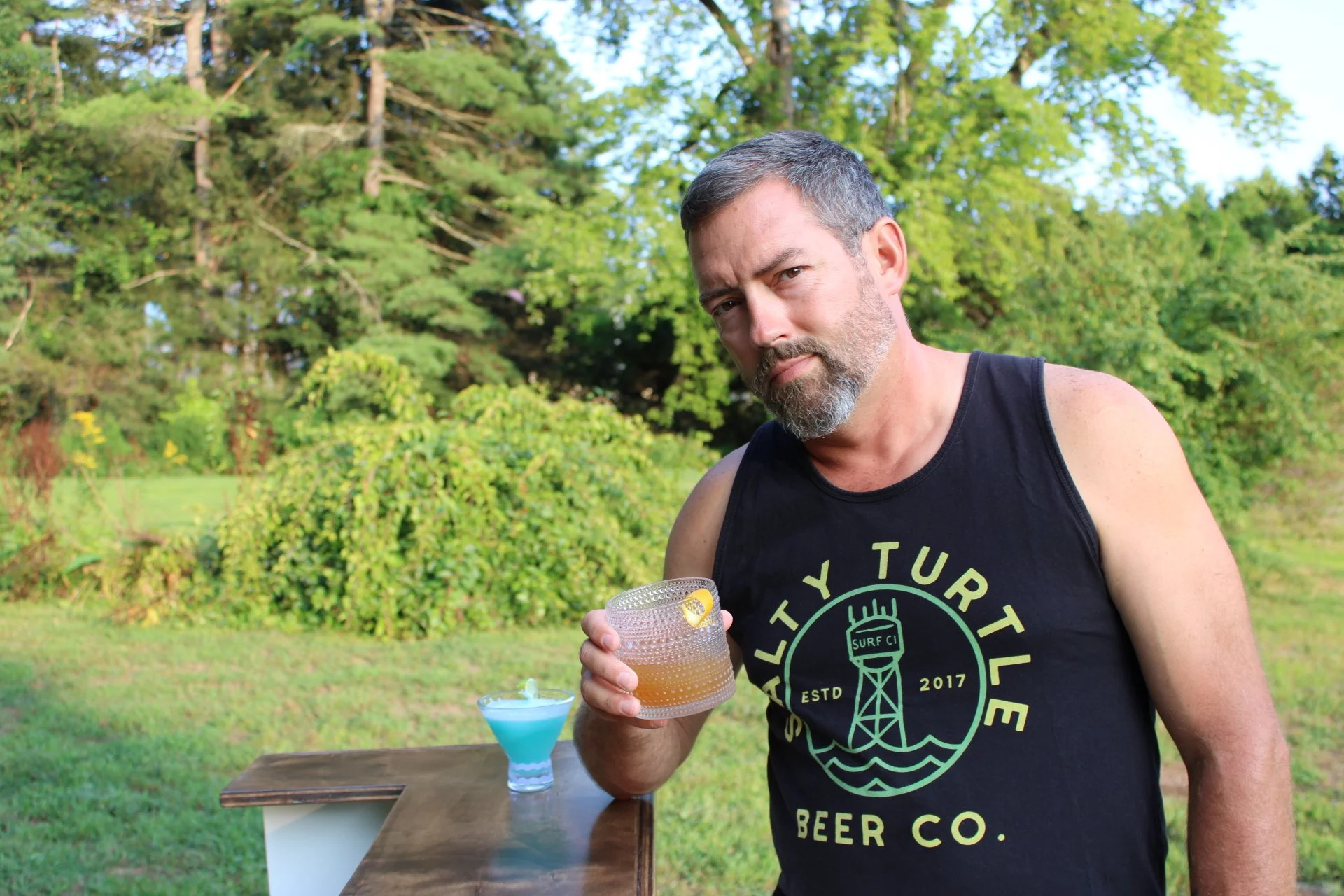 A man with gray hair and beard wearing a black tank top with green text and a graphic stands outdoors holding a drink in a textured glass. The background features green trees and bushes, with a blue cocktail on a wooden surface nearby.