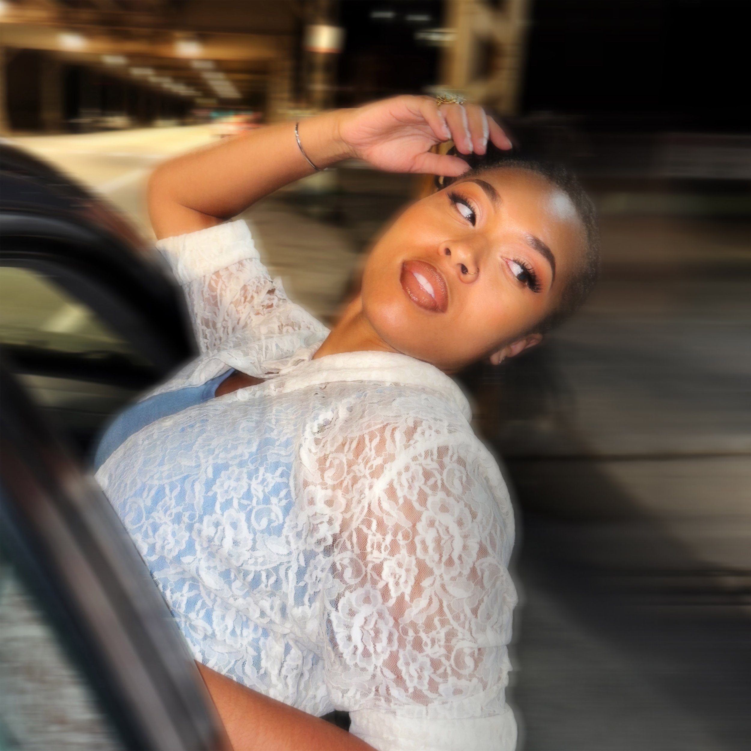 A woman with short hair, makeup, wearing a white lace top, sitting in a car with a blurred background, looking away from the camera.
