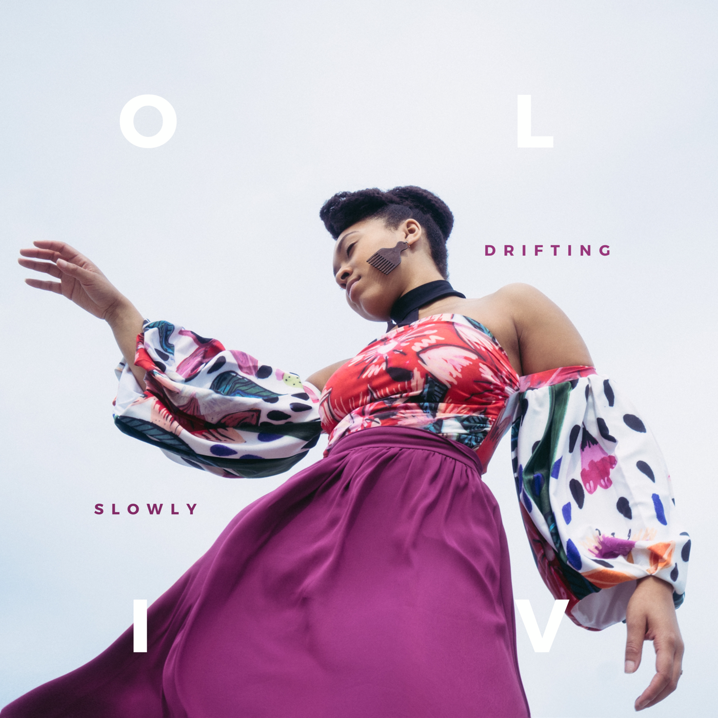 A woman with short dark hair wearing a colorful off-shoulder blouse with puffy sleeves and a purple skirt, posing outdoors against a cloudy sky. Oliv Blu slowly drifting cover art.