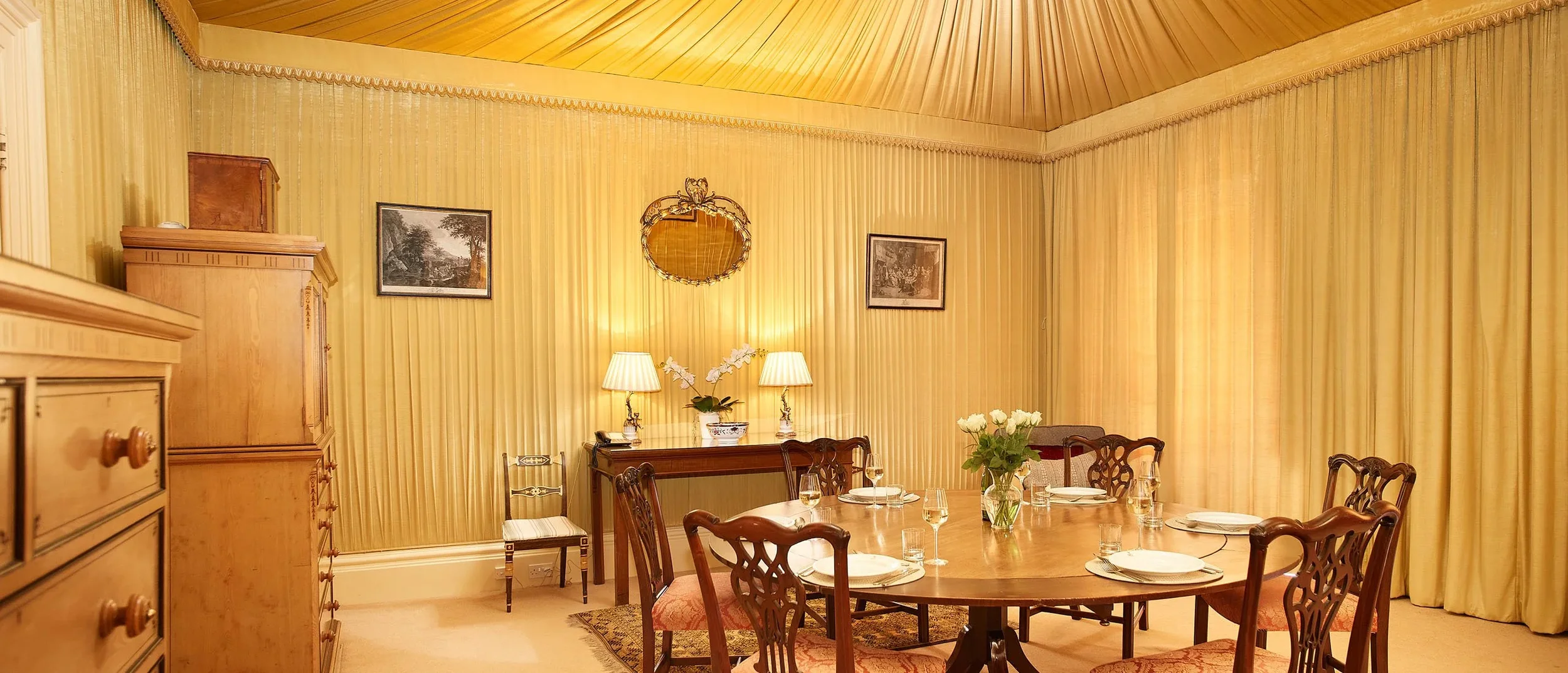 Living room with teal walls, wooden window frames, shelves with vintage equipment, framed pictures, a vase with white flowers, green patterned armchairs, dark wood furniture, and a ceiling light.