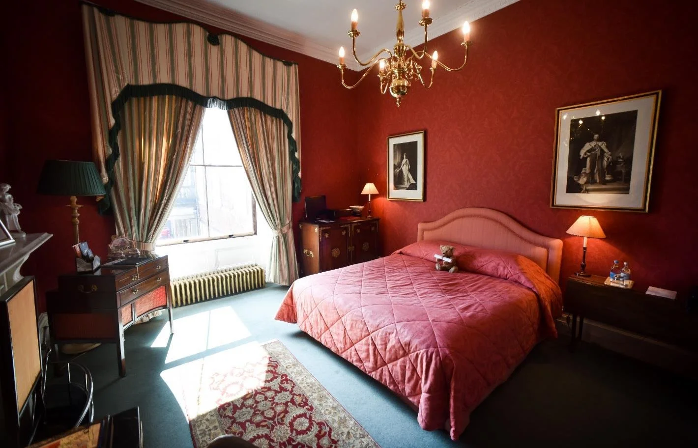 Bright bathroom with light blue walls, built-in bathtub, patterned curtains, a window with a scenic view, a small table with toiletries, and a wooden cabinet with a lamp on top.