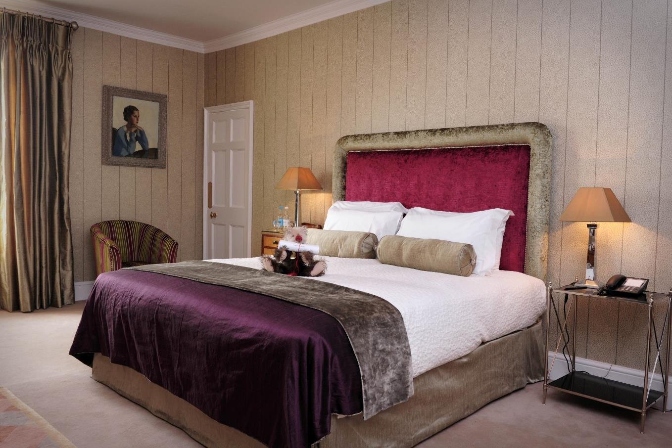 A bedroom with a purple accent wall, a double bed with religious-themed bedding, and a tufted headboard. There are two nightstands with lamps, a full-length mirror, a seating area with a pink velvet bench, framed artwork on the walls, and chandeliers hanging from the ceiling.