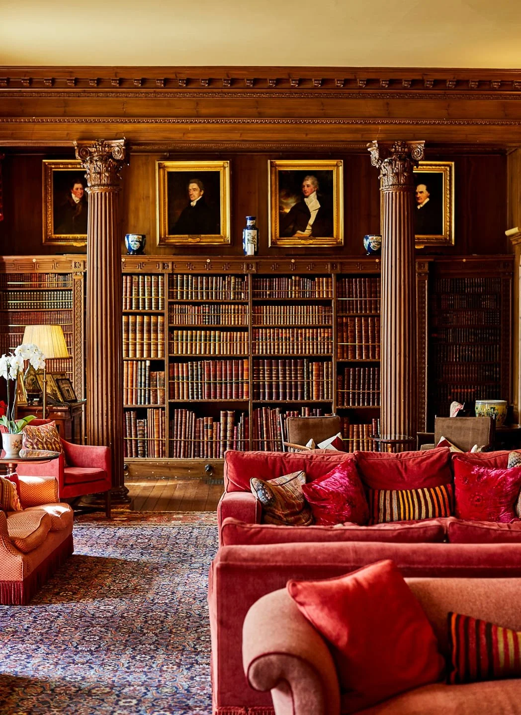 A cozy living room with large windows, wooden furniture, a blue sofa with a fur throw, armchairs, a fireplace, and decorative curtains and artwork.
