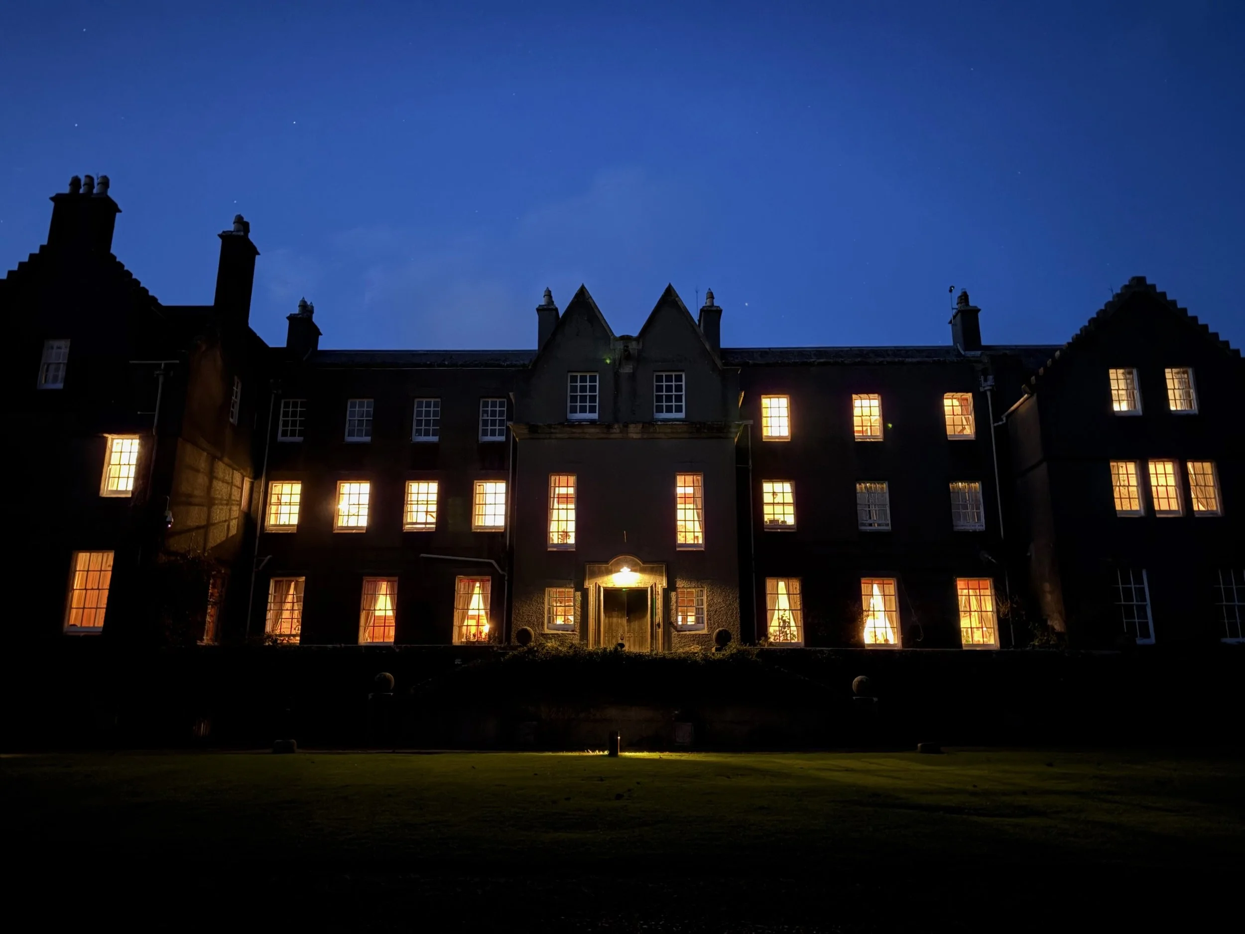 Large, multi-story house with a front porch, surrounded by lush greenery and trees, under a partly cloudy sky.