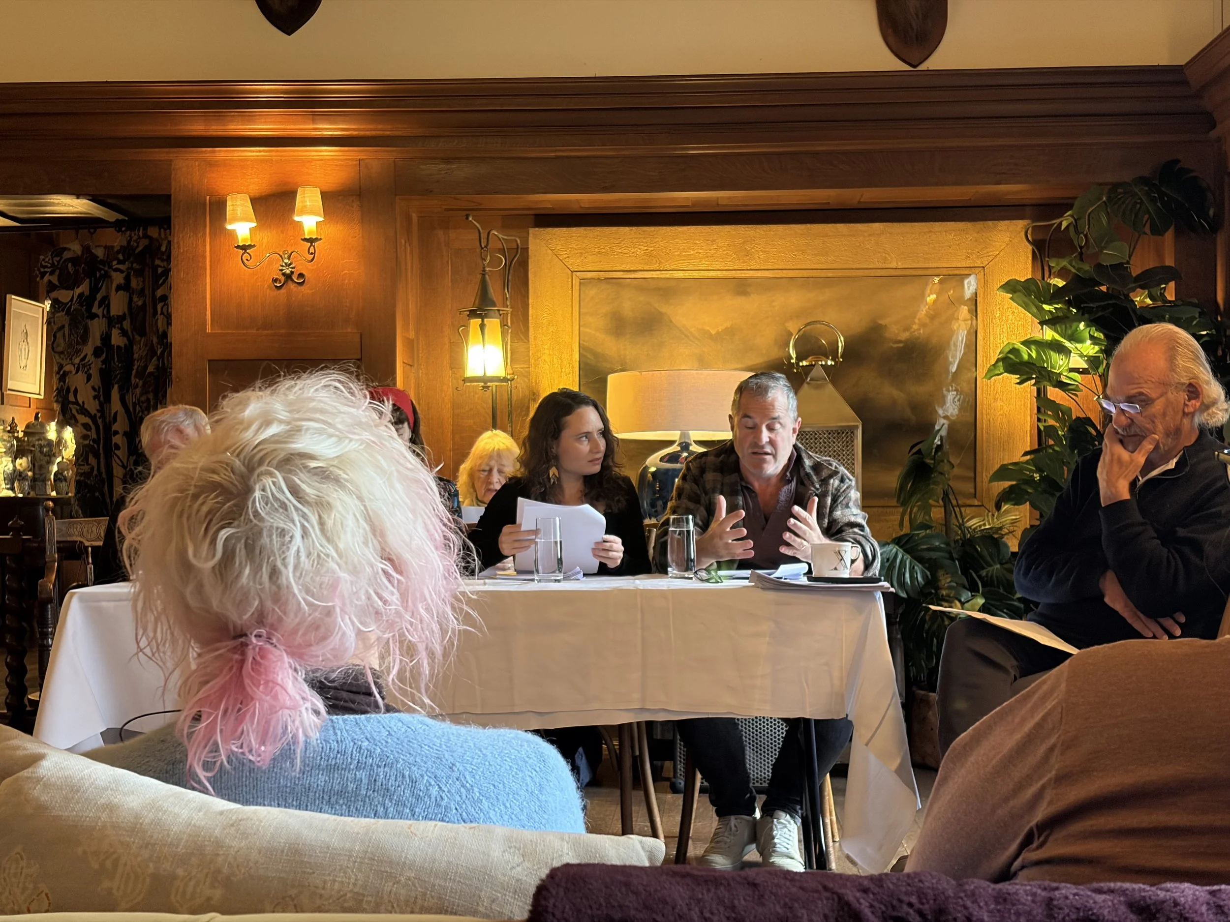 A woman sitting on a chair on a stage with a black background, holding a piece of paper in one hand, with her eyes closed and her other hand on her forehead, appearing stressed or overwhelmed.
