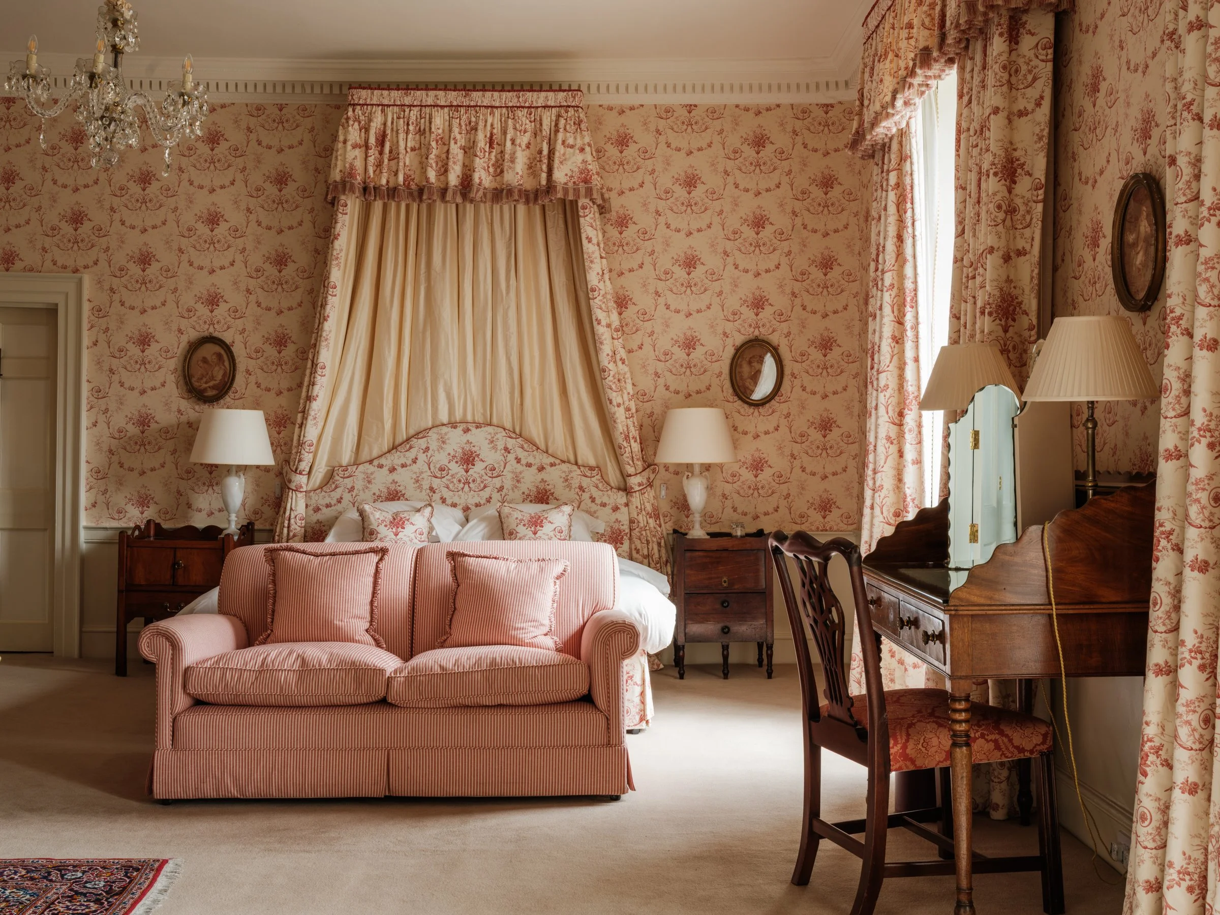 Interior of a vintage-style bedroom with green curtains, antique wooden furniture, a chandelier, and floral artwork on the wall.