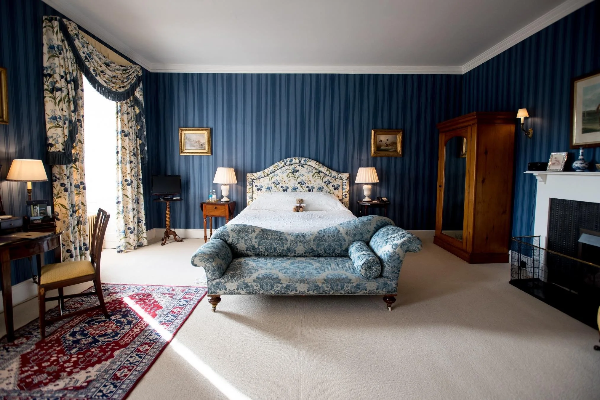 A bedroom with a large window with green curtains, a painted headboard, two matching bedside tables with lamps, a beige armchair with a purple pillow, and framed artwork on the wall. The bed has purple and blue pillows and a blue quilt.