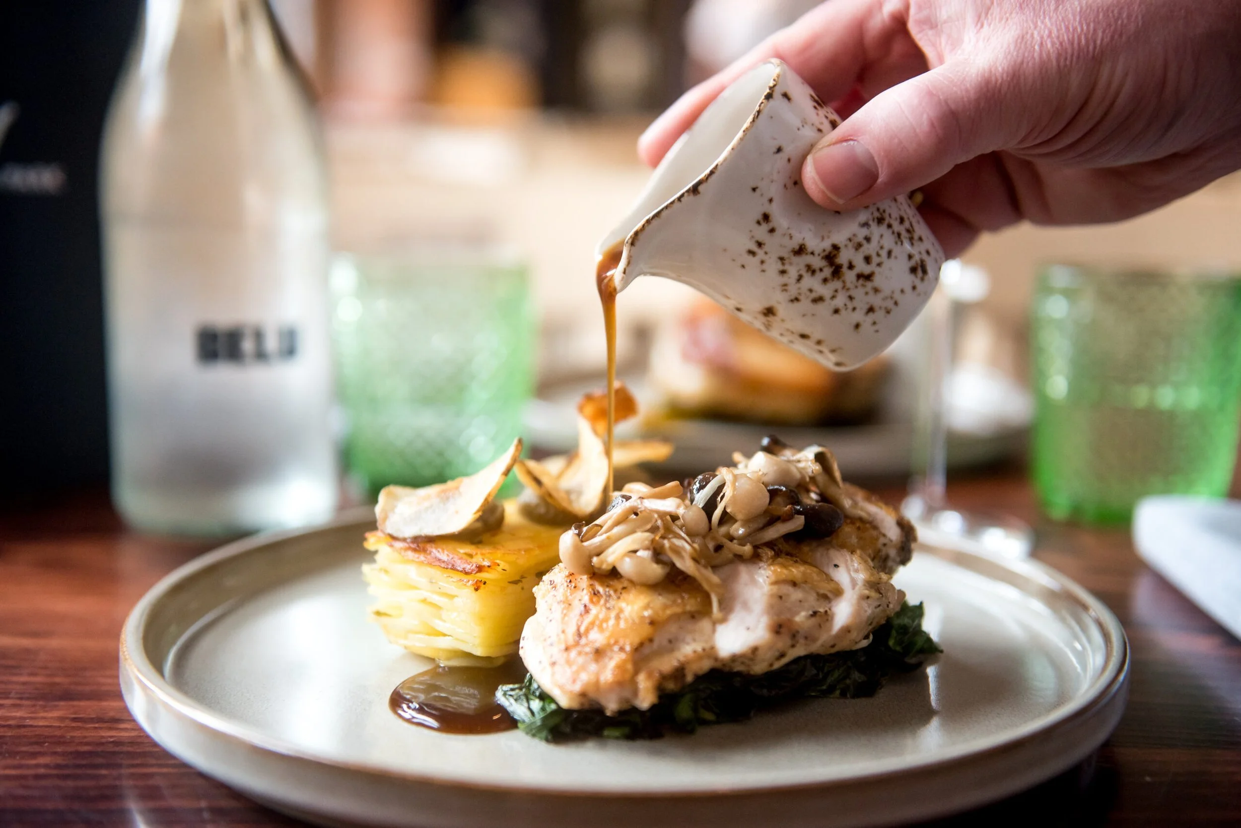 People sitting at a picnic table outdoors in a garden during daytime, enjoying food. In the background, a man and a woman are walking up a grassy hill lined with trees and bushes.