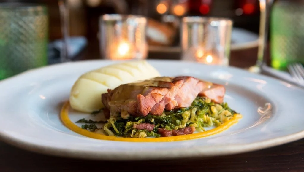 A gourmet salad with microgreens, watermelon, and carrots, garnished with colorful sauces, served on a green plate. A bowl of purple microgreens and a set of silverware are also visible on a dark cloth background.