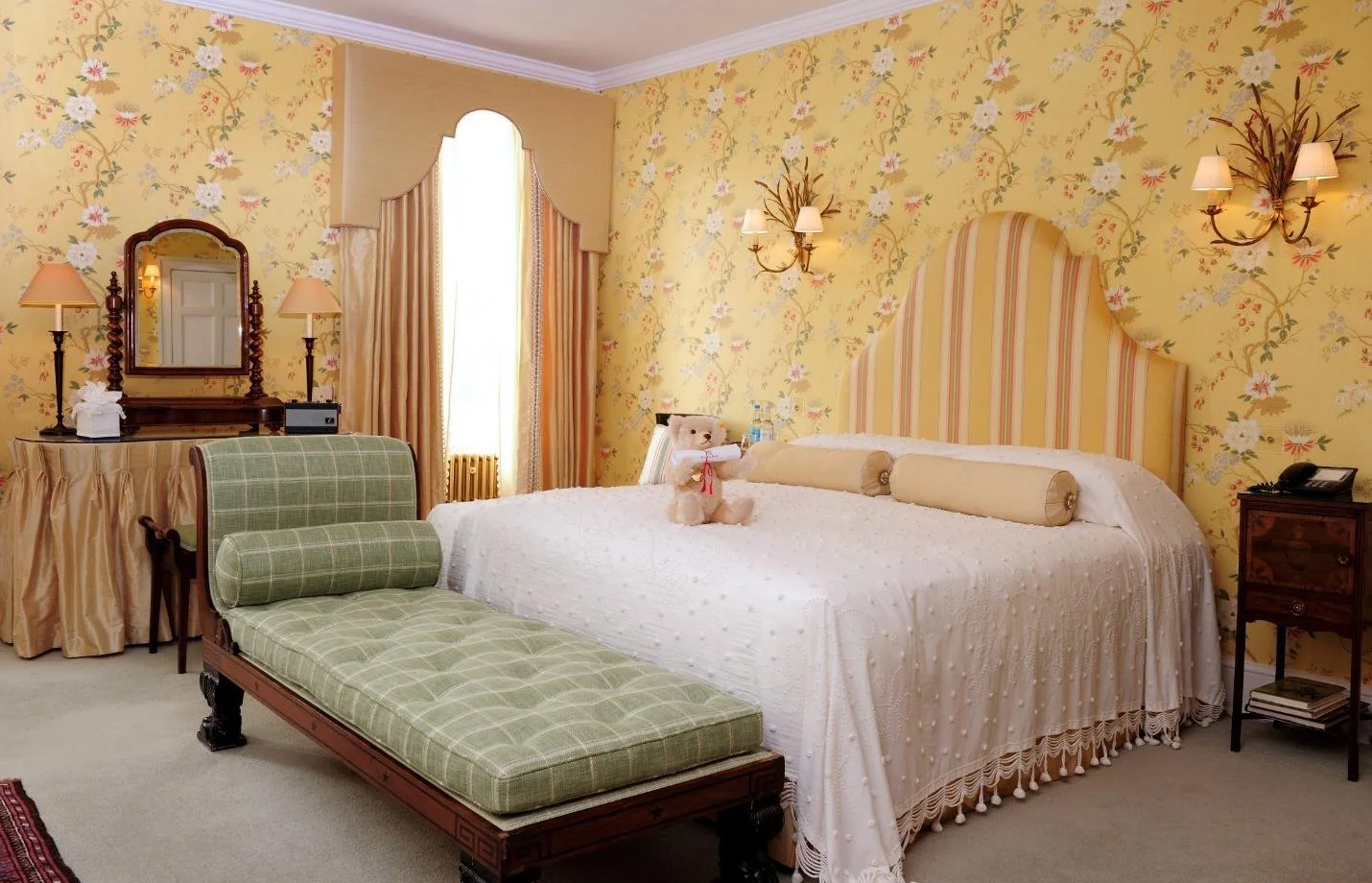 A cozy bedroom with a large bed, vintage furniture, and a window with curtains letting in natural light. Decor includes framed artwork, a patterned rug, and a fireplace