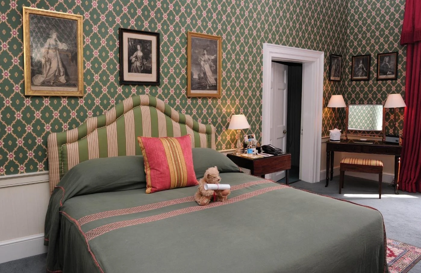 Elegant bedroom with light blue walls, white fireplace, and vintage furniture including a white bed, a white vanity, and a wooden chest of drawers, decorated with framed artwork, floral arrangements, and plush seating.
