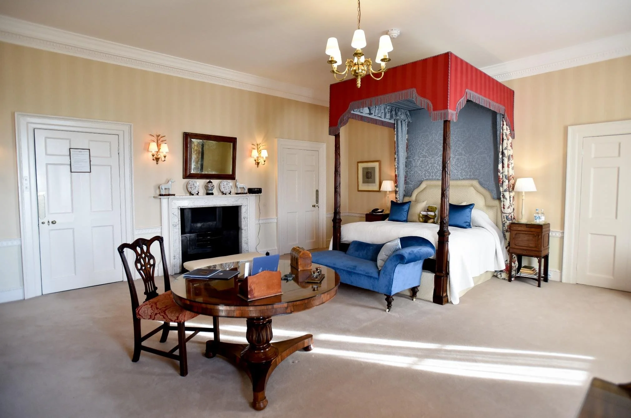 A vintage bathroom with white tile walls accented with decorative blue tile border, a white bathtub with a towel, a small white sink, a large mirror with ornate frame, hanging Moroccan-style lanterns, and various green potted plants on a small wooden table.