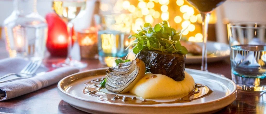 Elegant dining table set for a formal event with white tablecloths, tall candles, glassware, silverware, and flower centerpieces in a warmly lit room.