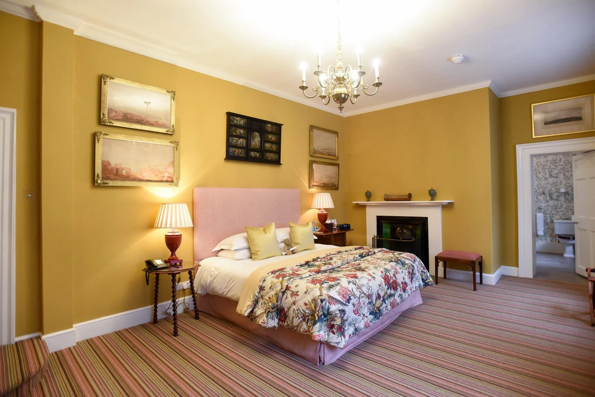 A vintage bedroom with a wrought iron four-poster bed, a floral dresser with a bouquet, a window with floral curtains, antique chairs, and in-room bathtub near a fireplace.