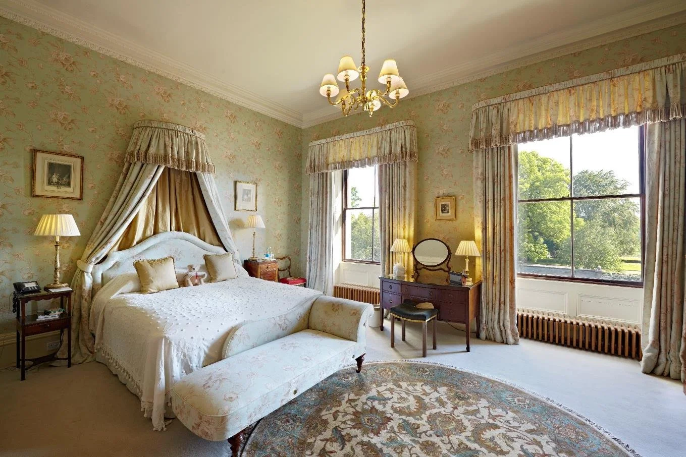 Luxury hotel room with separate bathtub, green velvet chaise lounge at the foot of a king-sized four-poster bed, and elegant decor including wall mirrors, framed pictures, and a gold-framed mirror.