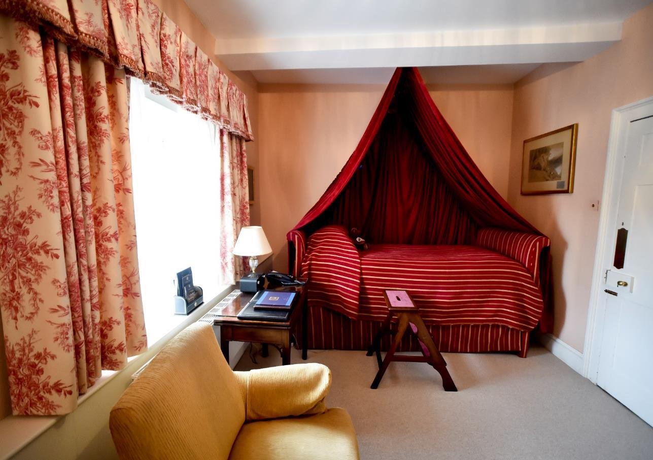 A bedroom with two brass beds, a colorful floral and bird-patterned wallpaper behind them, a large window with red curtains, a blue and orange patterned rug, and a sitting area with dark blue chairs and orange pillows.