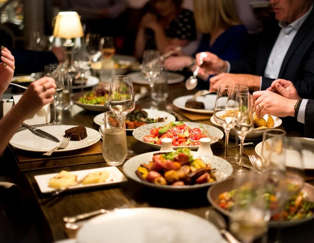 People socializing and laughing in a dimly lit room with a pool table, large windows, and framed artwork on the wall.
