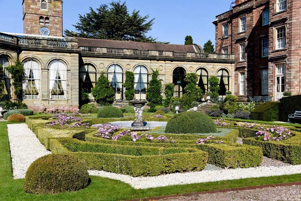 Aerial view of a historic estate surrounded by lush green trees, featuring a maze garden, several buildings, and well-maintained gardens.