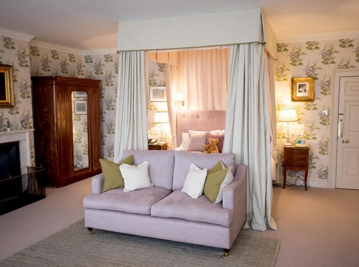 A bedroom with a large teal velvet headboard, floral wall art, a chandelier, and about four floral paintings, with nightstands on each side holding lamps and flowers, a window with green curtains, and a bed with a colorful quilt and pillows.