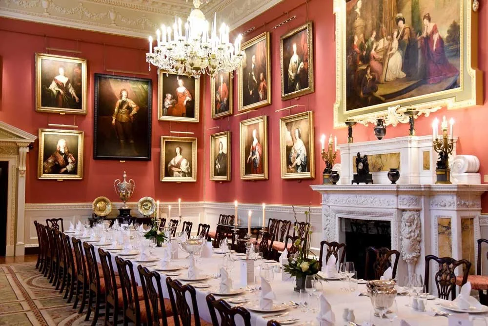 Elegant dining room with a large floral centerpiece, surrounded by candles, glasses, and tableware. The room features ornate crown molding, framed portraits on the walls, and a chandelier hanging from the ceiling.