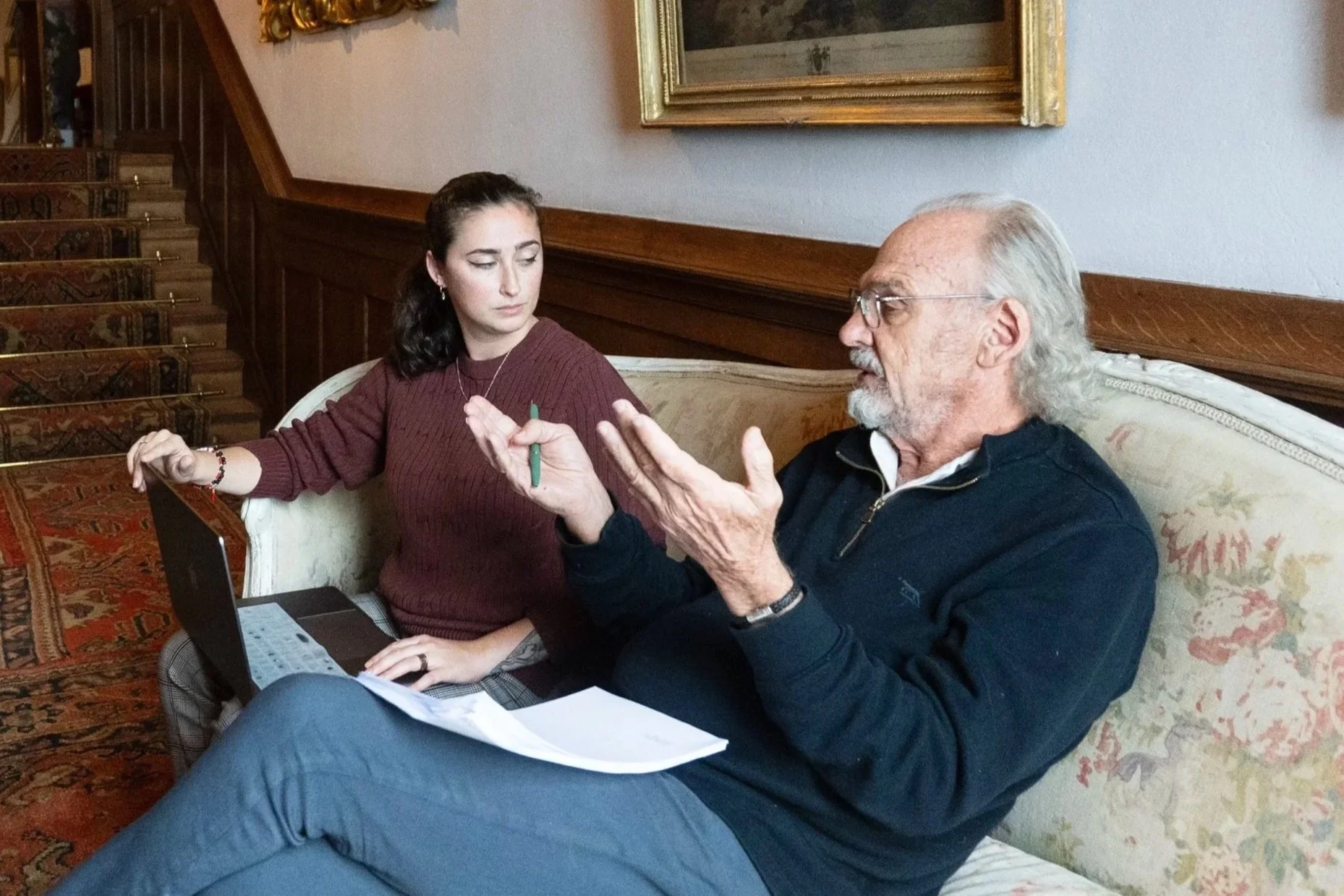 Christopher Lockhart, a man wearing glasses and a pink blazer sitting on a director's chair, speaking into a microphone on stage with a red curtain backdrop