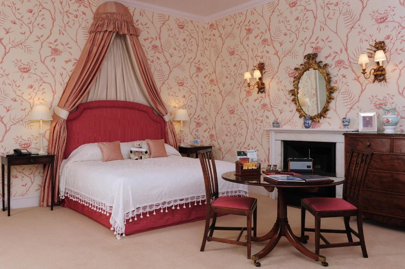 Cozy bedroom with a large window, a white bed with decorative pillows, teal accent wall with a round sunburst mirror, framed art, and side tables with lamps and floral arrangements.