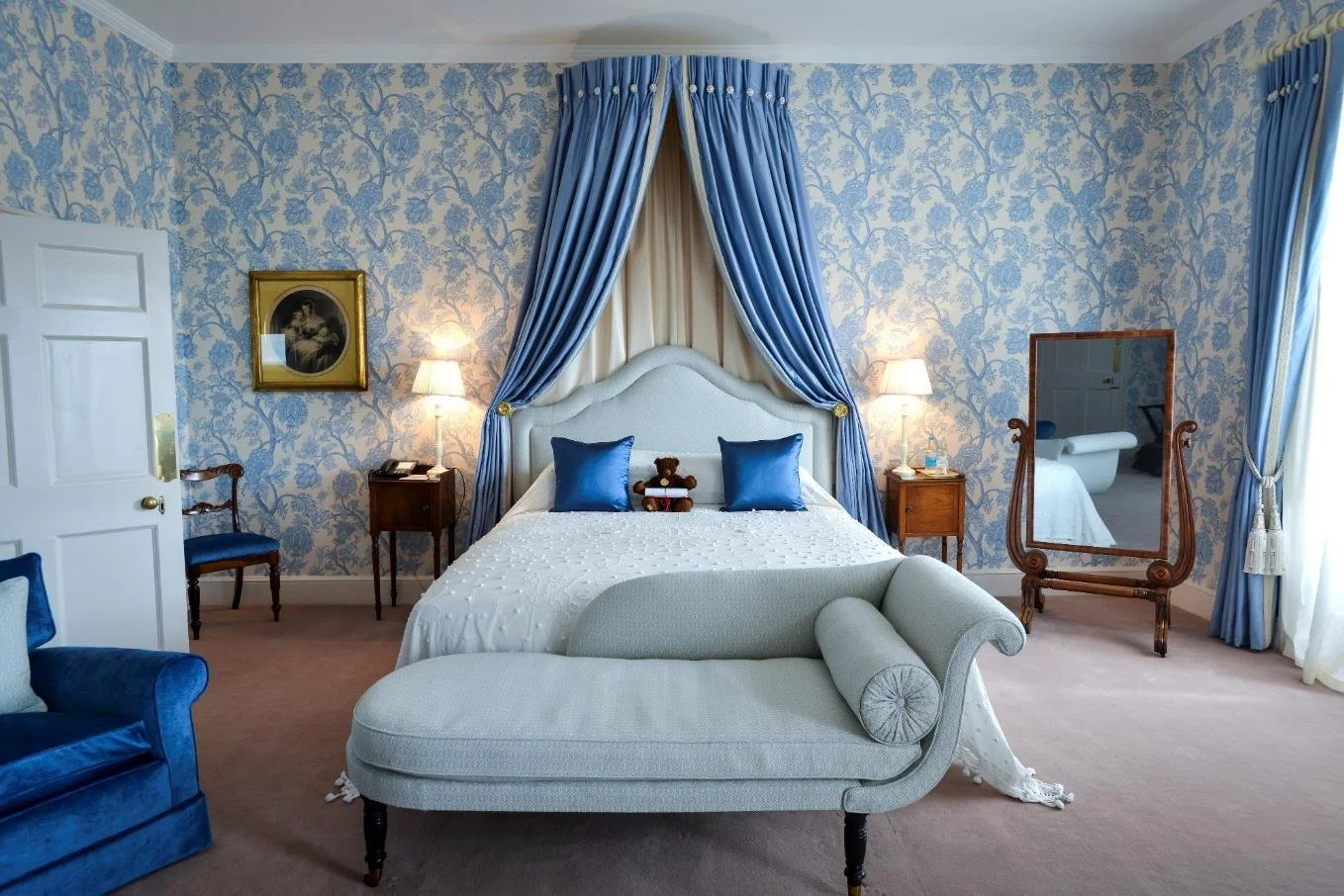 A bedroom with navy blue walls, floral curtains, a window with natural light, a bed with white and blue bedding, a rattan headboard, two table lamps, and Asian-inspired wall hangings.