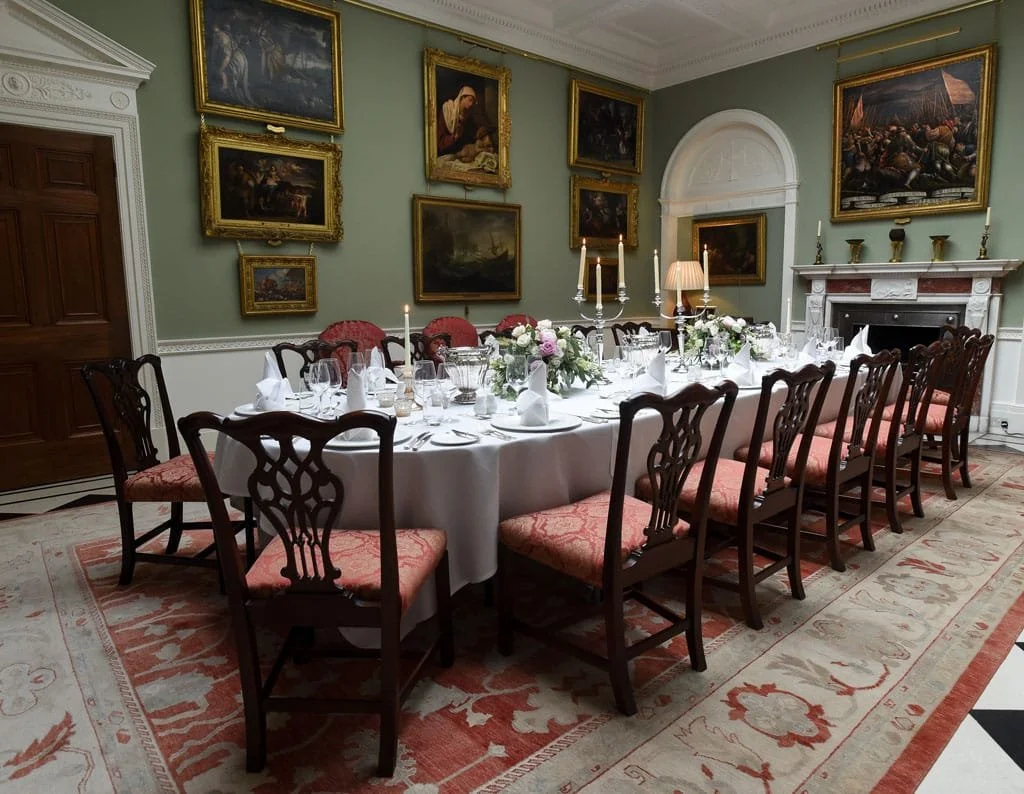 A beautifully decorated dining and living room with wooden paneling, large windows with curtains, a fireplace, decorative rugs, a dining table set with white linens and floral centerpieces, chairs, and mounted deer heads on the wall.