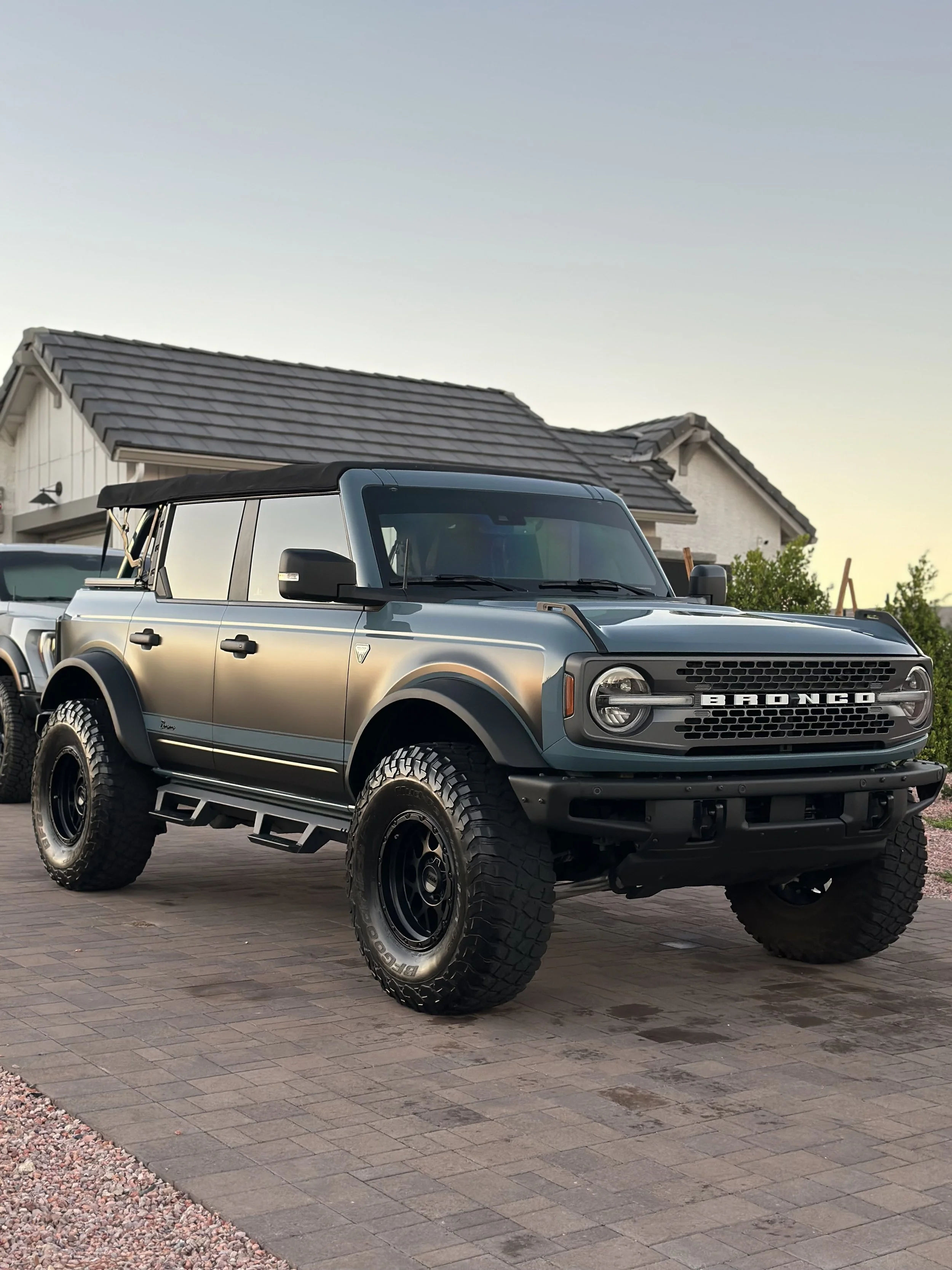 A Ford Bronco SUV parked on a brick driveway in front of a house with a shingled roof, during sunset.