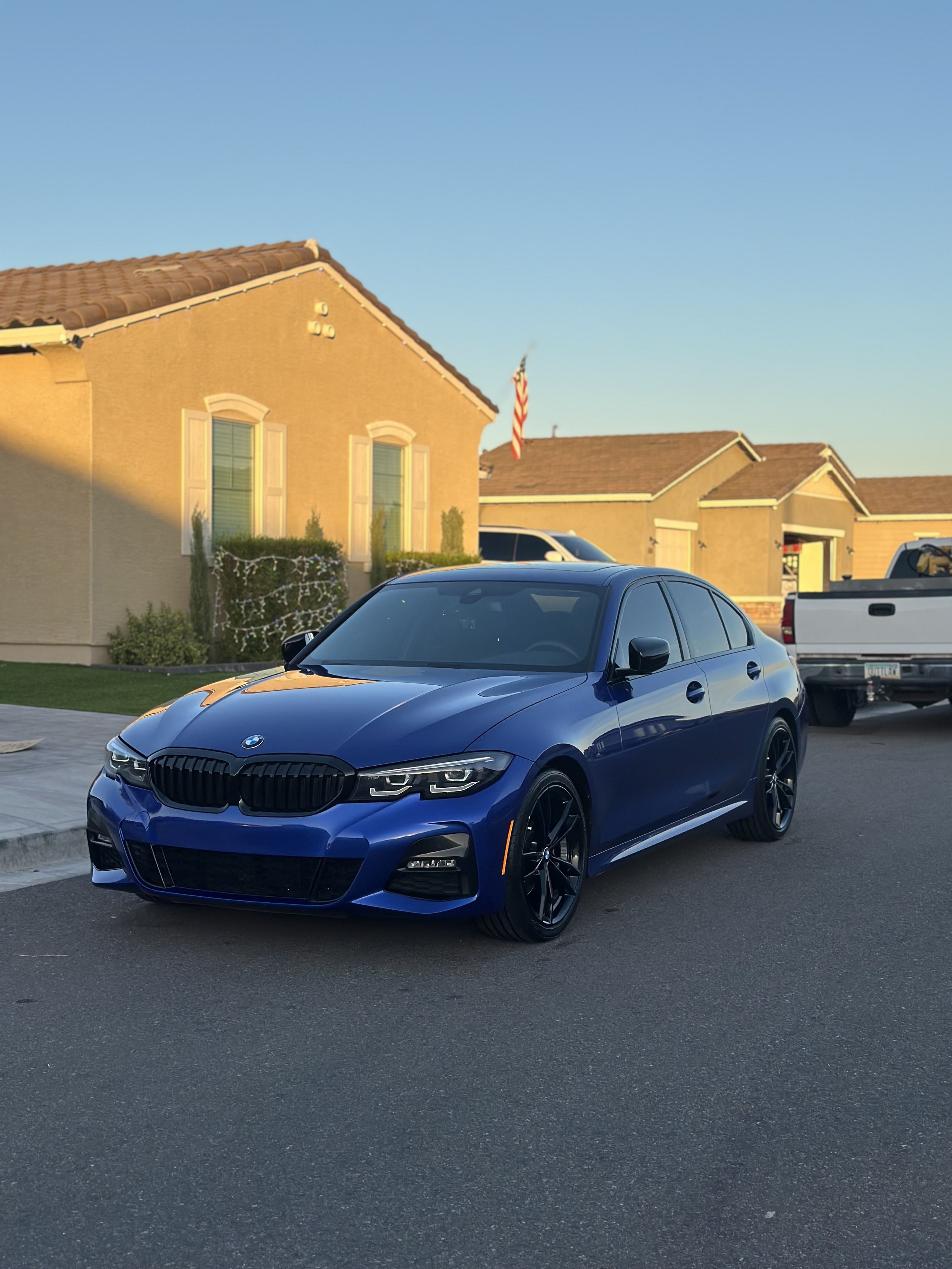 A blue BMW sedan parked on a residential street during sunset with houses and an American flag in the background.