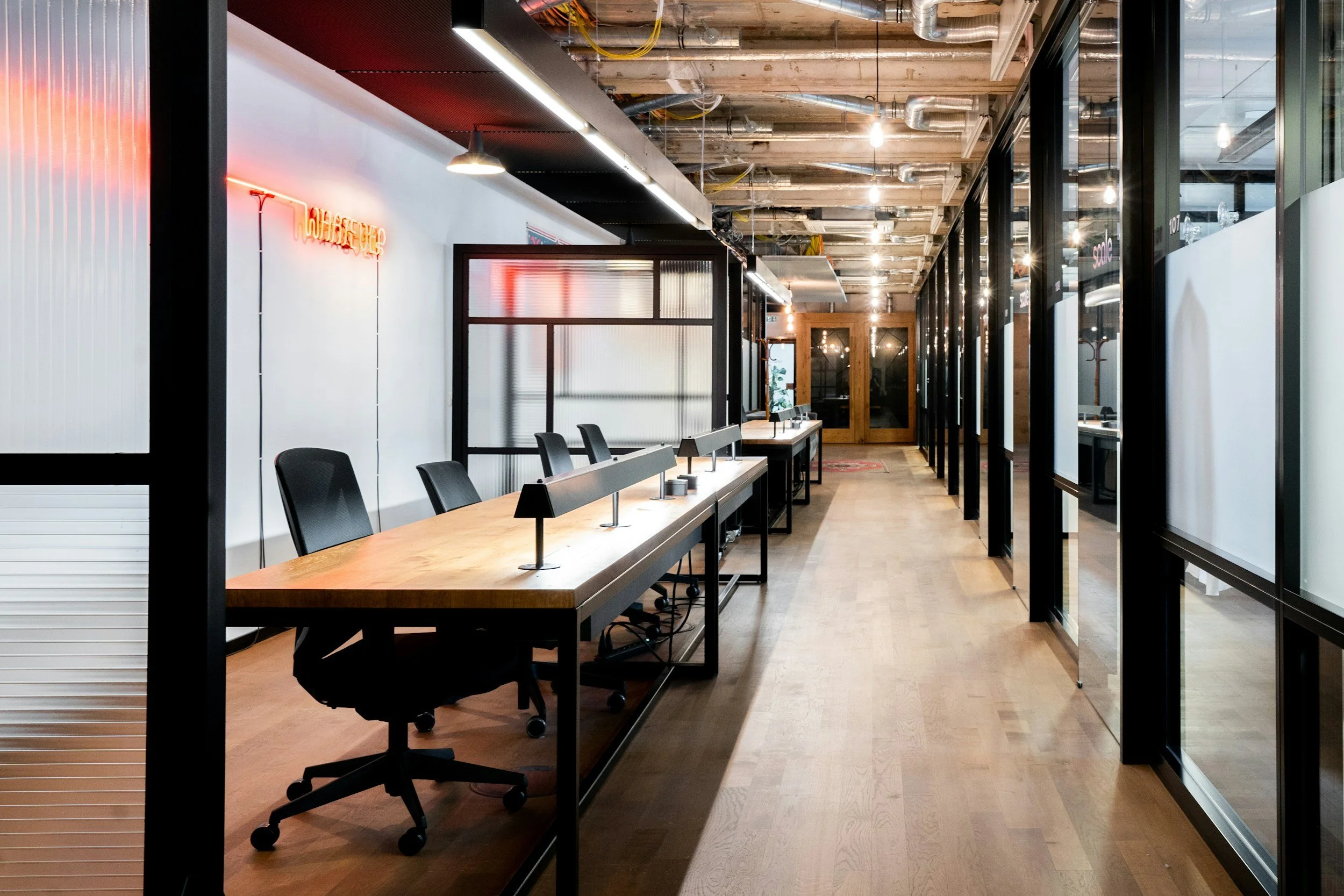 Modern office space with a long wooden conference table, black office chairs, glass partition walls, and exposed ceiling with lighting fixtures.