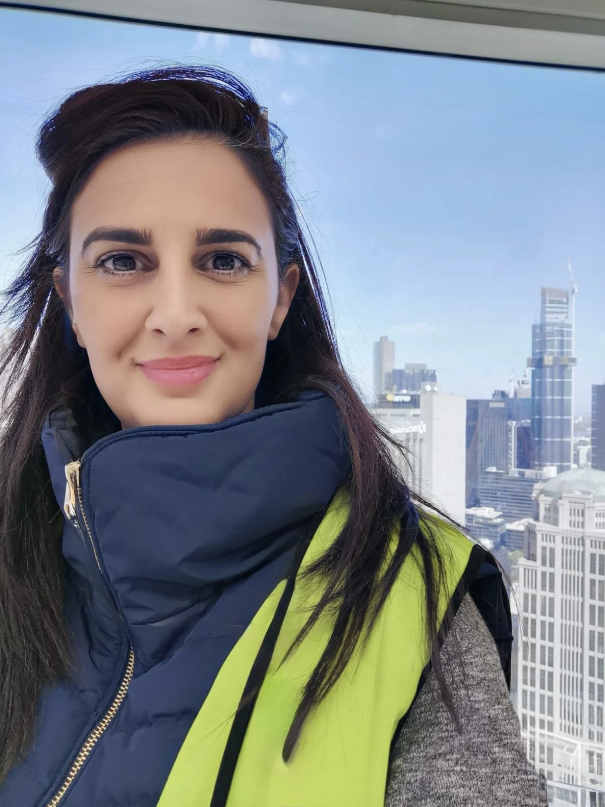 A woman with brown hair and blue eyes taking a selfie in a tall building, wearing a yellow safety vest and a dark blue jacket, with a city skyline in the background.