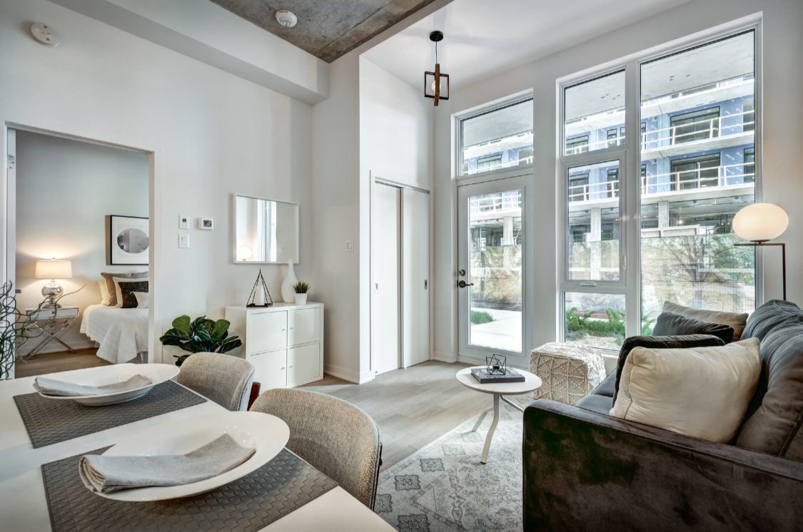 Modern living room with large windows, a gray sofa, white cushions, a small round coffee table, and a patterned area rug. There is a floor lamp, a white dresser with decor, and a view of an apartment building outside.